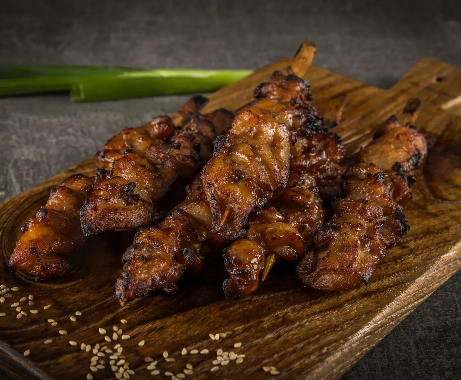 Mehrere goldbraun gegrillte Fleischspieße mit leicht karamellisierter Oberfläche liegen auf einem Holzbrett, bestreut mit Sesamkörnern. Im Hintergrund ist eine grüne Lauchzwiebel zu sehen.