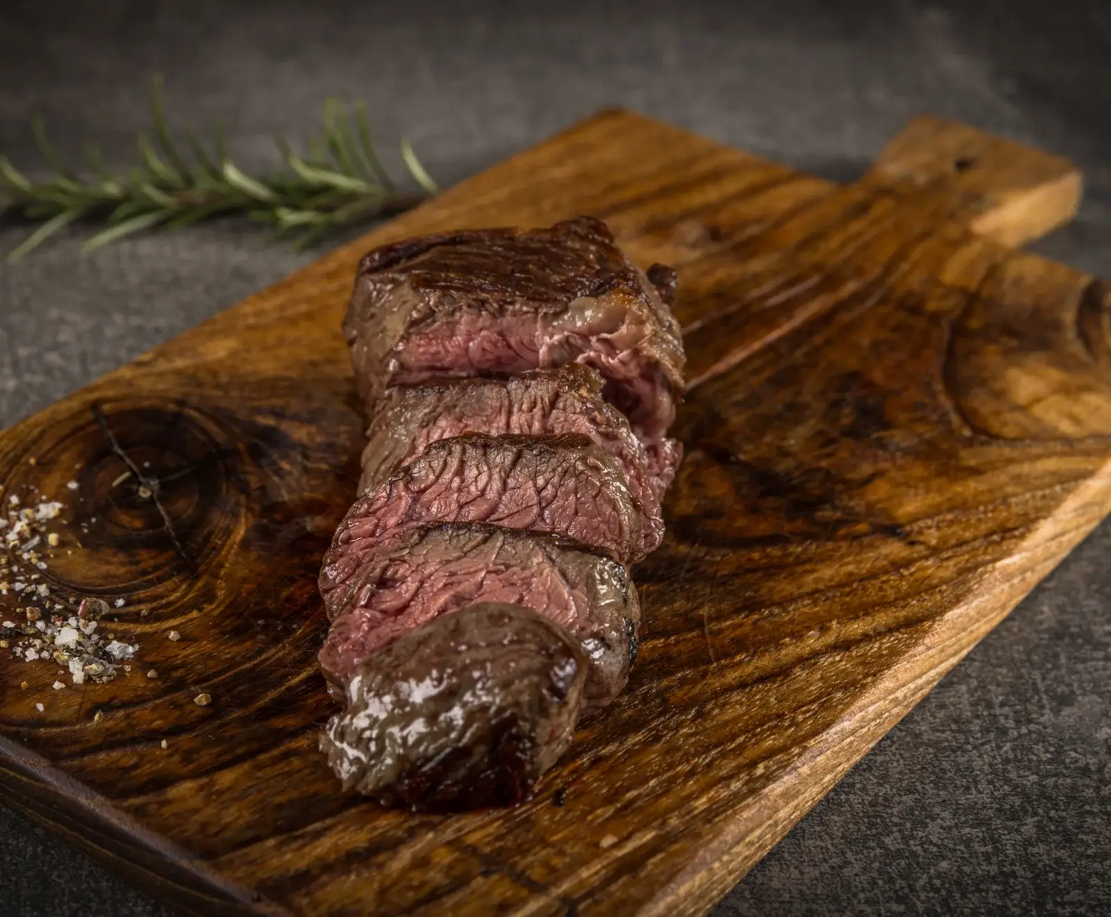 Ein perfekt gegartes, in Scheiben geschnittenes Steak liegt auf einem rustikalen Holzbrett. Daneben sind grobes Salz und Pfeffer sowie ein Rosmarinzweig zu sehen.