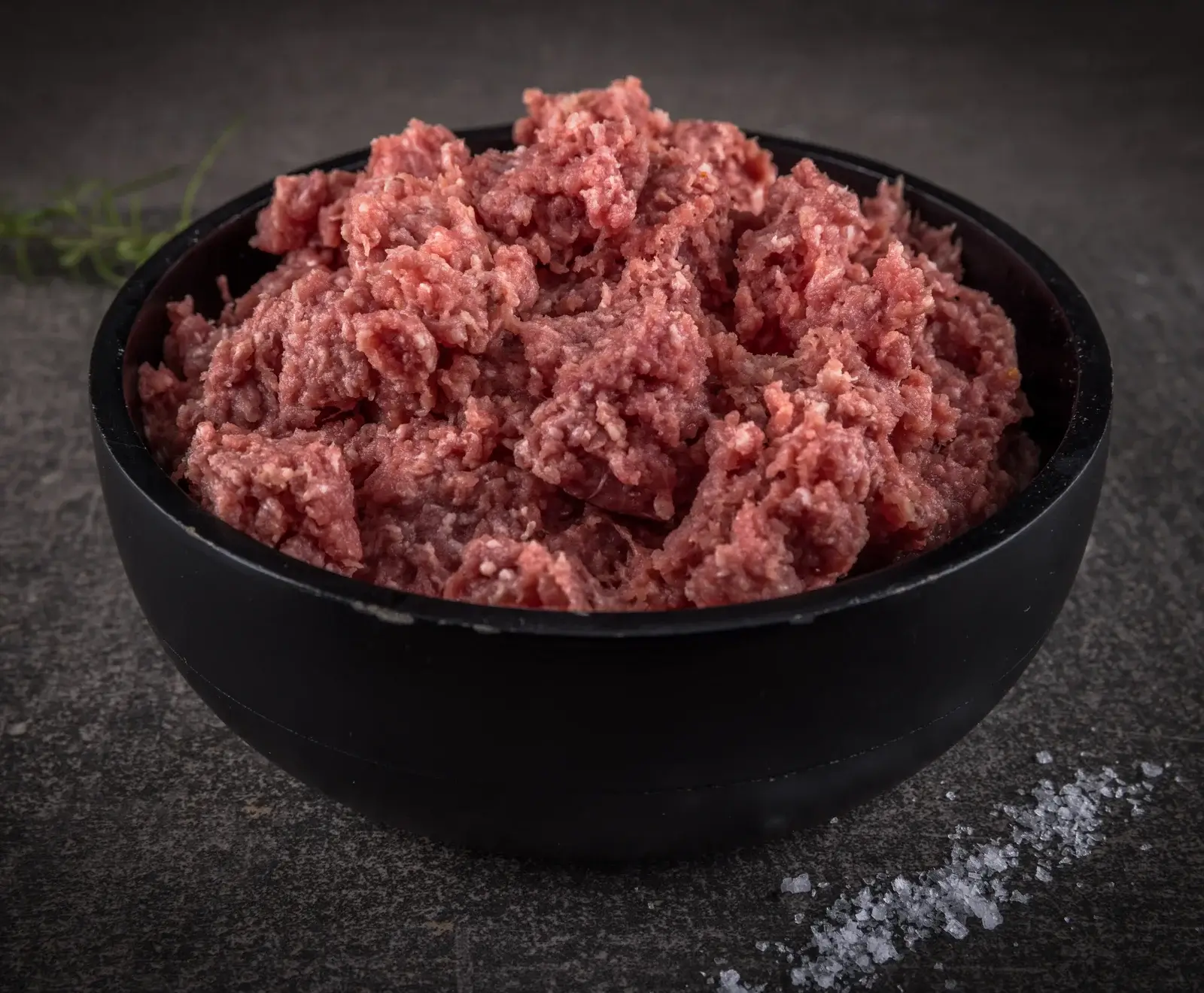 Eine schwarze Schale gefüllt mit frischem, rohem Hackfleisch auf einer dunklen, strukturierten Oberfläche. Im unscharfen Hintergrund ist ein Rosmarinzweig, im Vordergrund grobes Salz zu sehen.