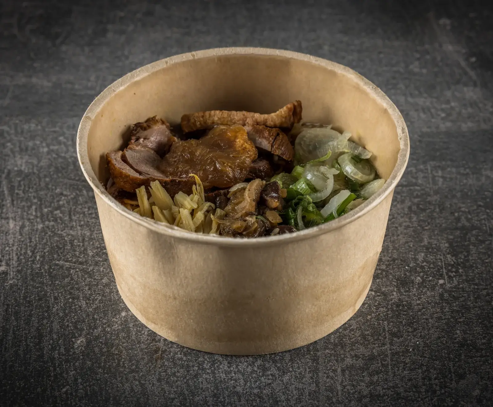 Eine braune Pappschale mit einem asiatischen Gericht. Es enthält geschnittenes Fleisch (vermutlich Ente oder Schweinebauch), geschmorte Pilze, eingelegtes Gemüse und frische Frühlingszwiebeln.