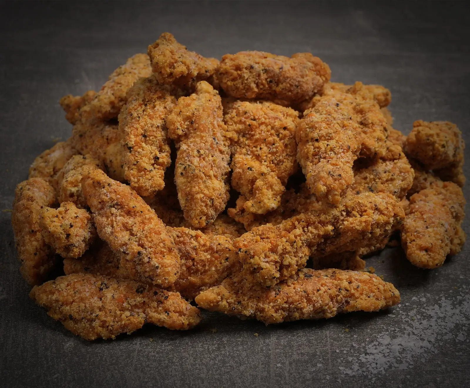 Ein Haufen gefrorener, panierter Hähnchen-Tender oder -Nuggets mit sichtbaren Gewürzen auf einer dunklen, grauen Oberfläche.