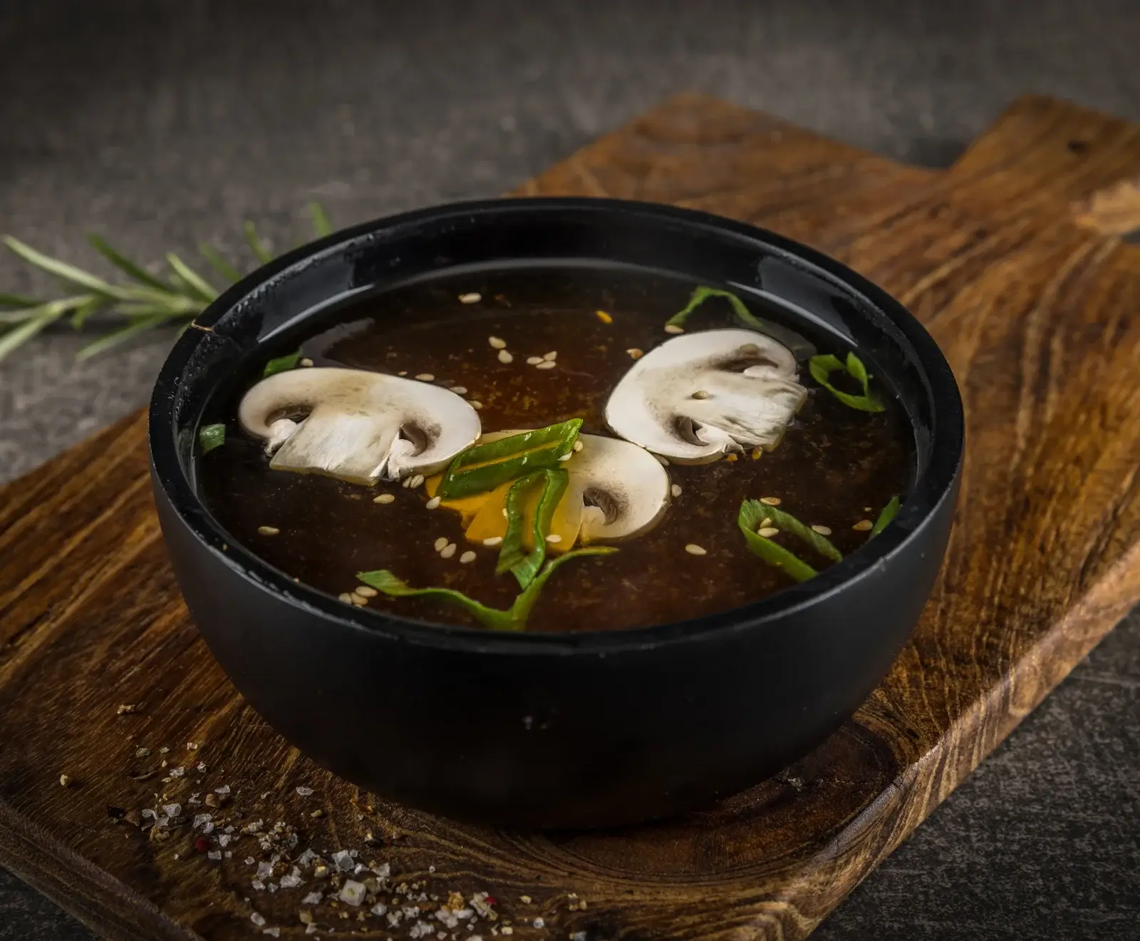 Eine schwarze Schale mit dunkler Pilzsuppe, garniert mit Champignonscheiben, Frühlingszwiebeln und Sesamsamen, steht auf einem rustikalen Holzbrett mit Gewürzen.