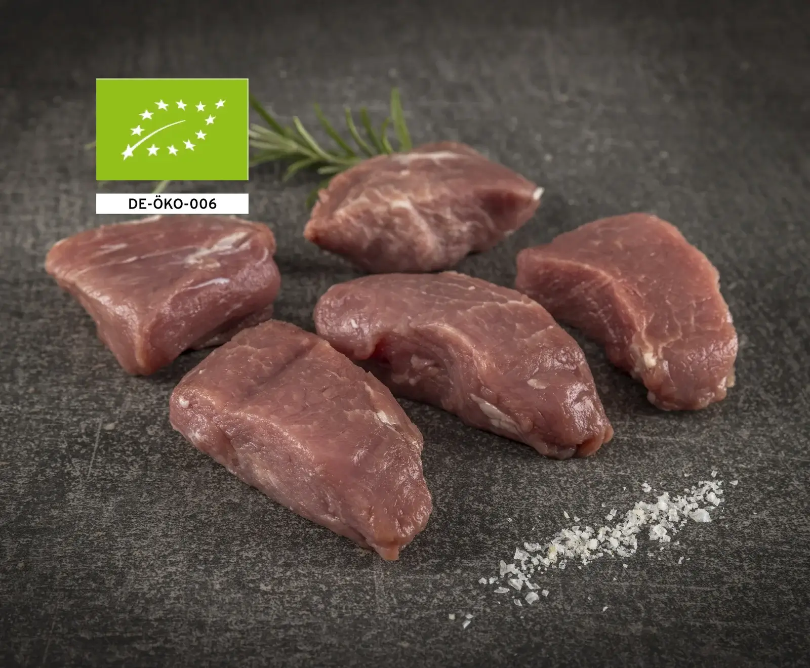 Mehrere rohe Bio-Rindersteaks auf einer dunklen Oberfläche mit Salz und Rosmarin garniert.