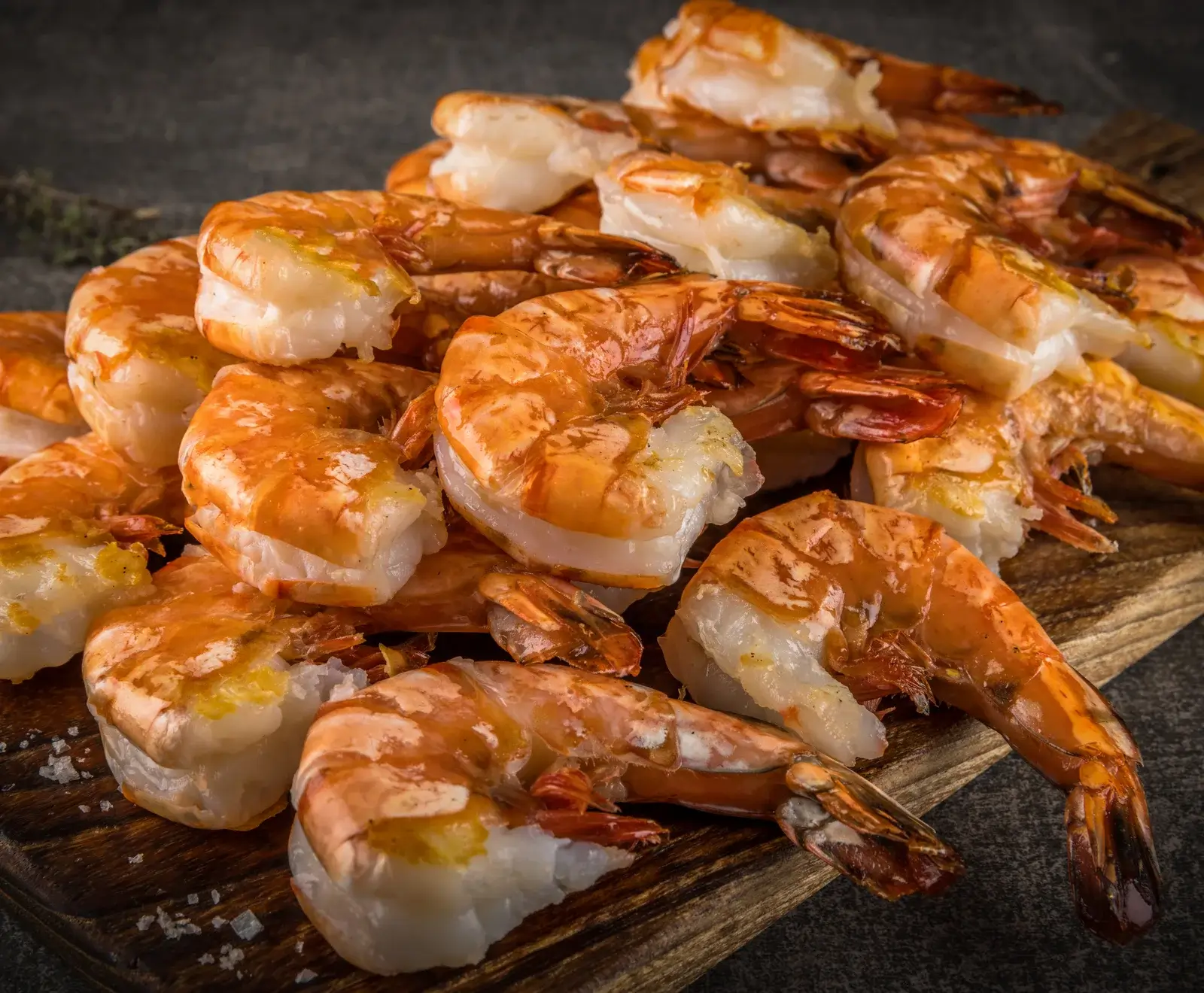 Ein Haufen gekochter, geschälter Garnelen mit Schwanz auf einem dunklen Holzbrett, mit einigen Salzkristallen und einem unscharfen dunklen Hintergrund.