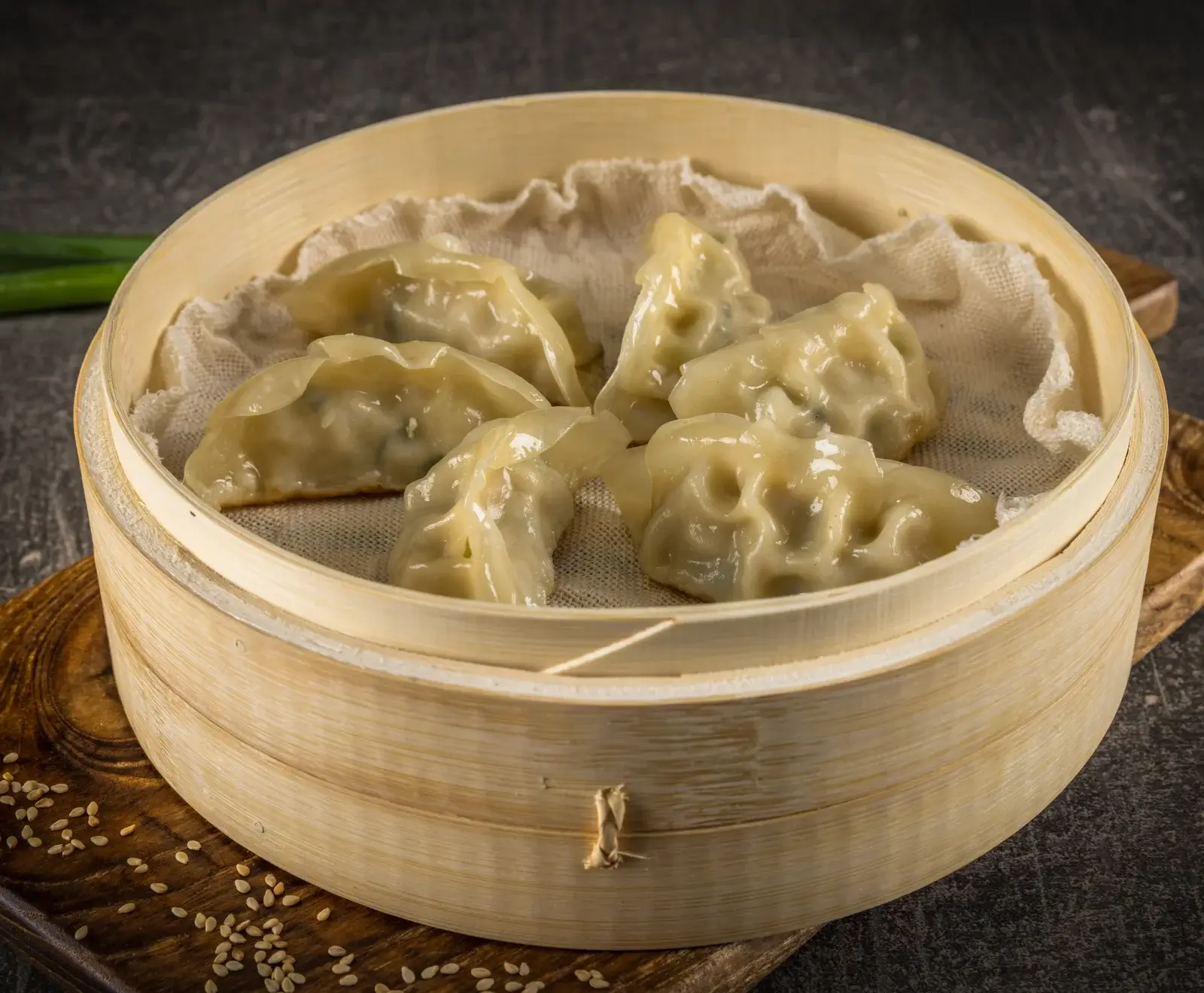 Mehrere glänzende, gedämpfte Teigtaschen (Gyoza/Jiaozi) liegen auf einem weißen Tuch in einem runden Bambusdämpfer. Im Hintergrund sind grüne Zwiebeln und Sesamsamen auf einem Holzbrett zu sehen.