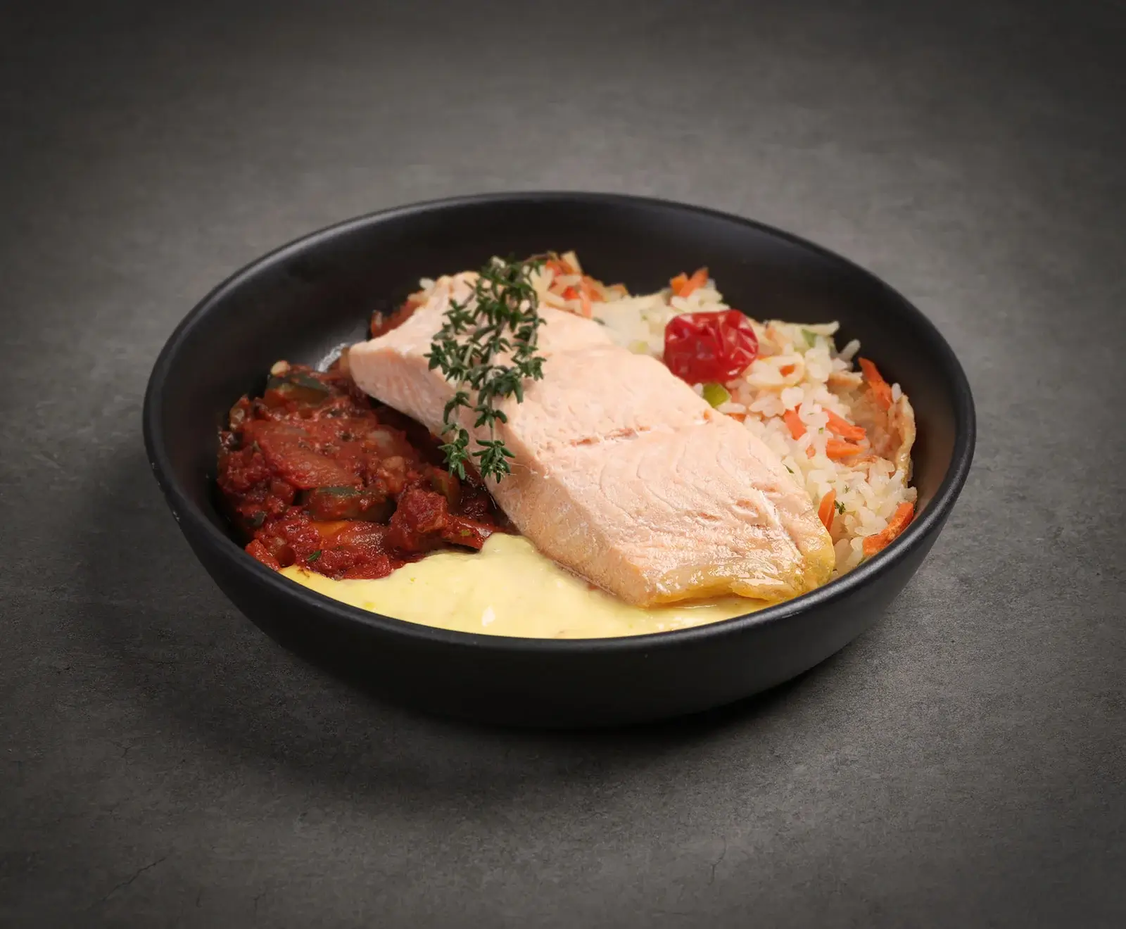 Ein gekochtes Lachsfilet mit Thymianzweig, serviert in einer schwarzen Schale. Dazu Ratatouille, eine helle cremige Sauce und Reis mit Karotten und Kirschtomate.