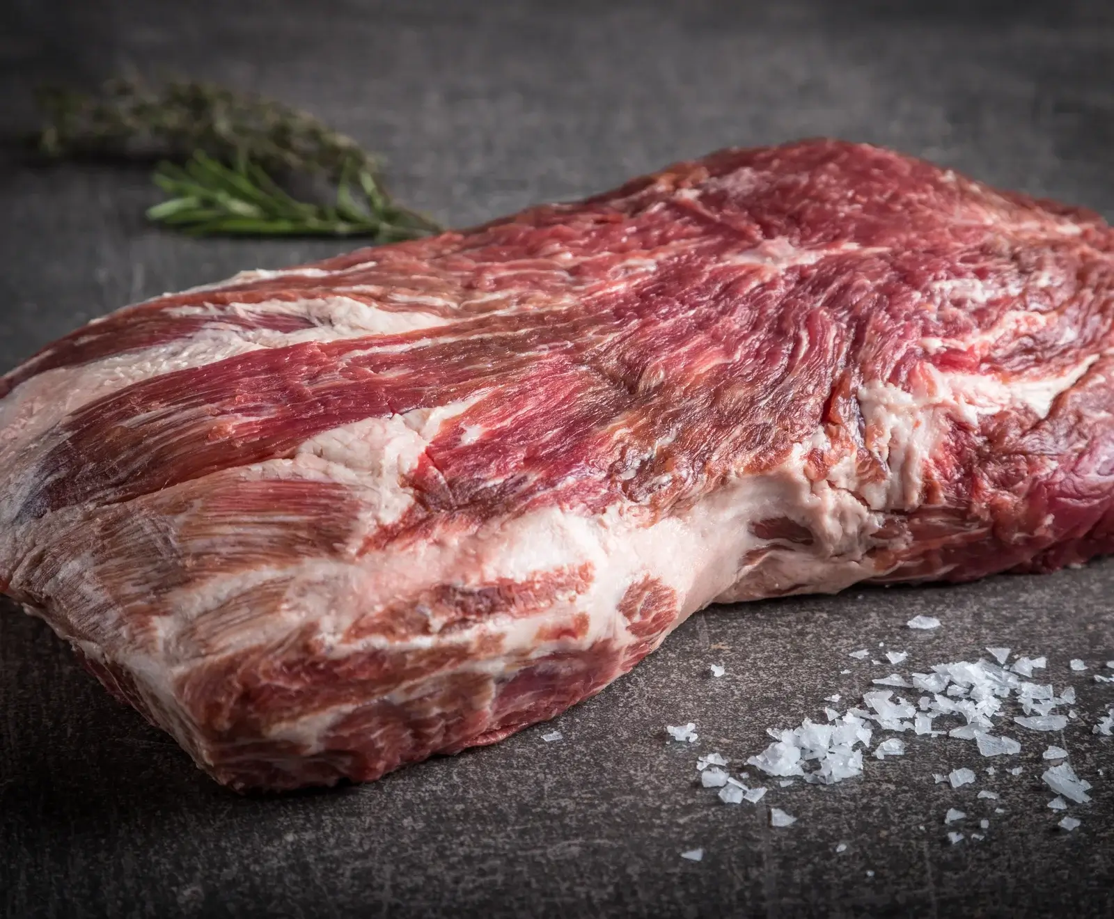 Rohes Rindfleisch, marmoriert mit Fett, auf grauem Hintergrund mit Salzkristallen und Rosmarin garniert.