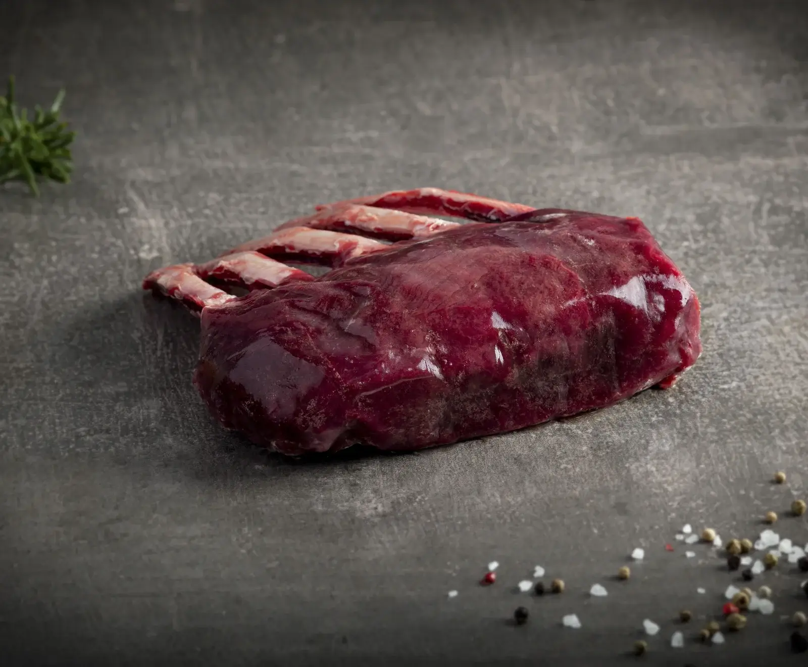 Ein rohes, dunkelrotes Stück Wildfleisch mit Knochen (Rehrücken) liegt auf dunklem Untergrund. Links oben ein Rosmarinzweig, rechts unten Pfefferkörner und grobes Salz.