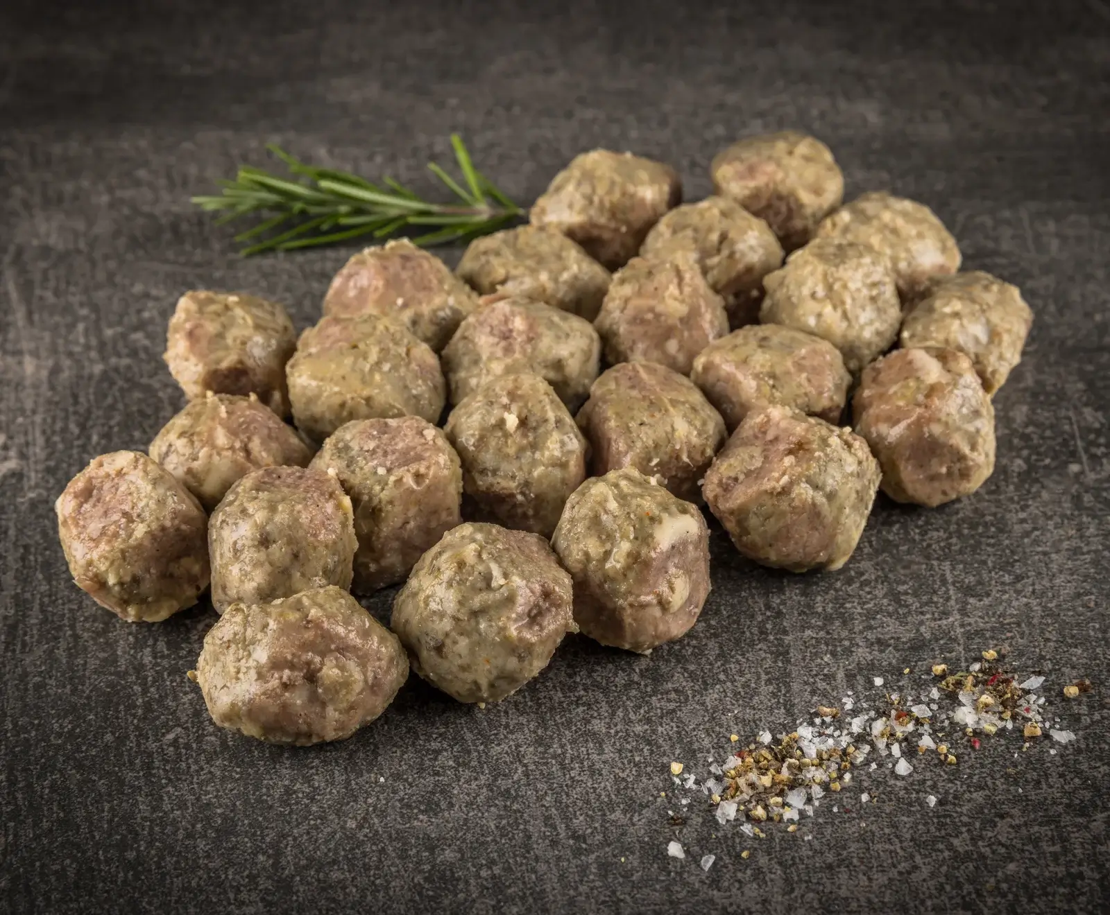 Ein Haufen gekochter, hellbrauner Fleischbällchen liegt auf einem dunklen, strukturierten Untergrund. Im Hintergrund ist ein Rosmarinzweig, im Vordergrund grobes Salz und Pfeffer.