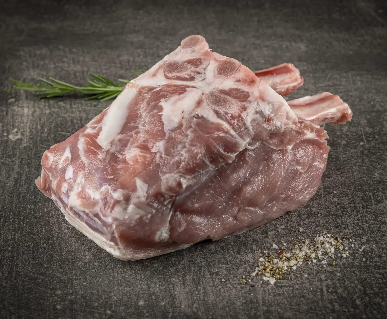Rohes Karree vom Schwein oder Lamm mit sichtbaren Rippenknochen, Fettmarmorierung, einem Rosmarinzweig und einer Prise Salz und Pfeffer auf dunklem, strukturiertem Hintergrund.