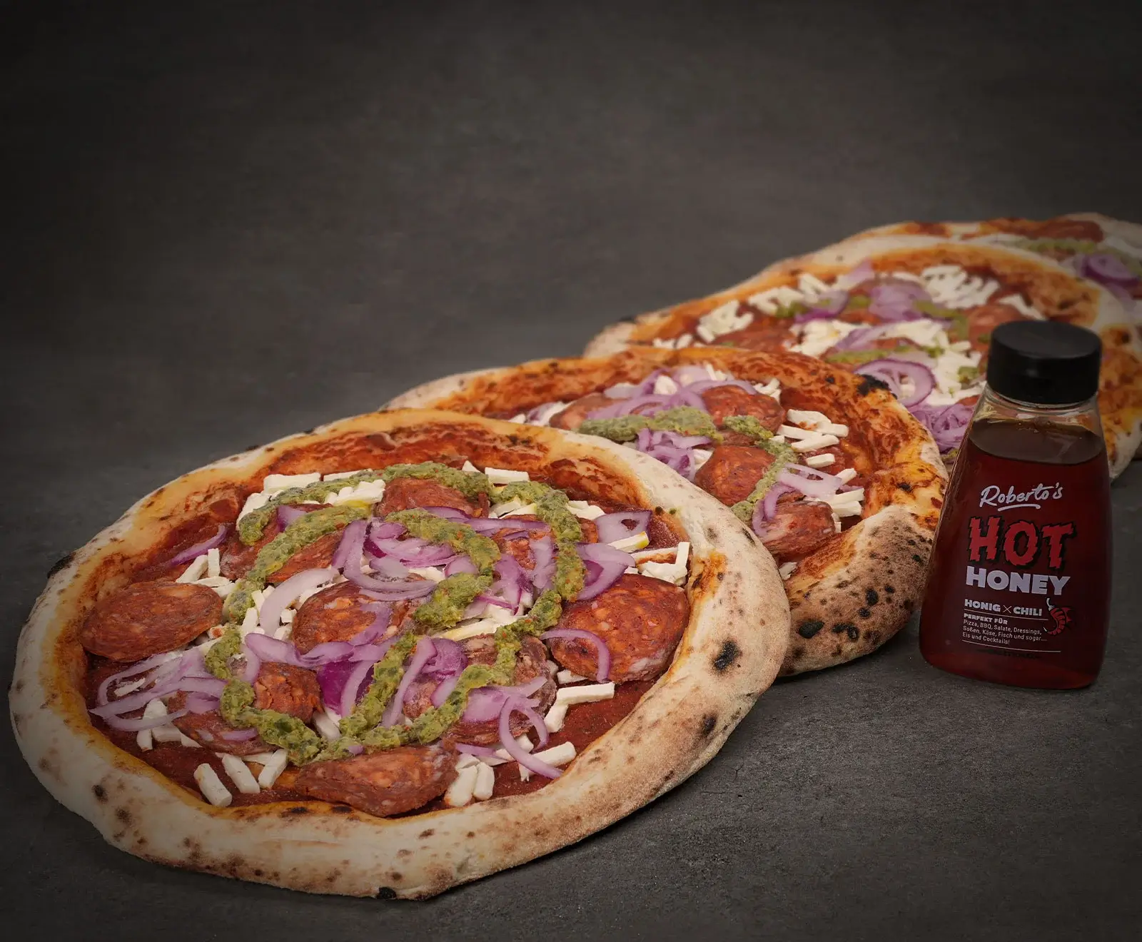 Drei ungebackene Pizzen mit Salami, roten Zwiebeln, Käse und grüner Soße auf dunklem Untergrund, daneben eine Flasche Roberto's Hot Honey Honig x Chili.