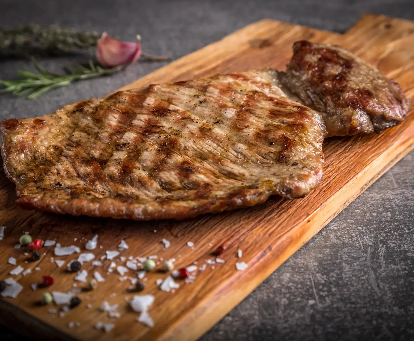 Zwei saftige, gegrillte Fleischstücke mit deutlichen Grillstreifen auf einem Holzschneidebrett, umgeben von grobem Salz, bunten Pfefferkörnern, Rosmarin und Knoblauch.