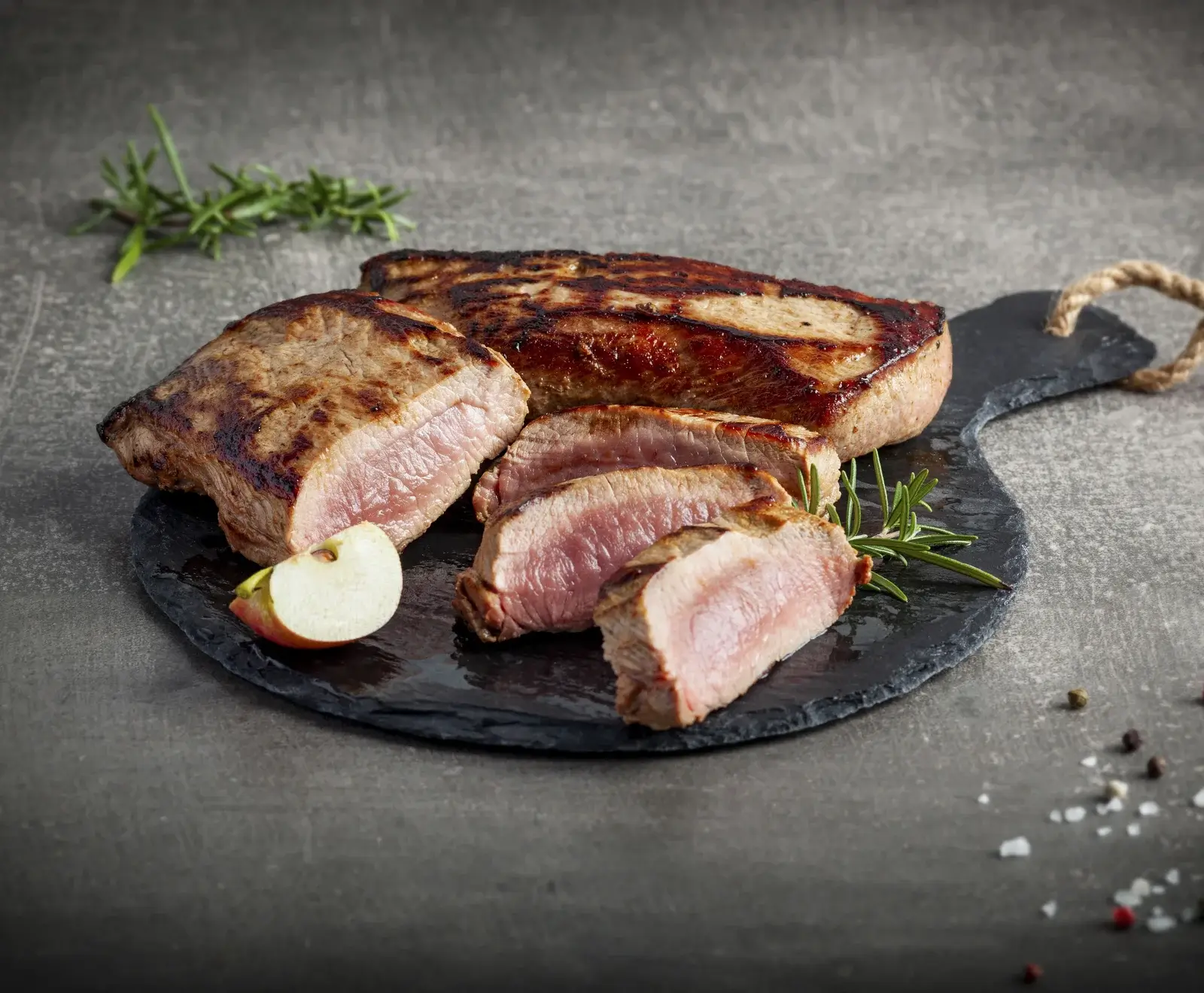 Ein aufgeschnittenes, medium gebratenes Stück Fleisch mit knuspriger Kruste auf einer schwarzen Schieferplatte. Dazu Rosmarin, ein Apfelstück, Pfefferkörner und Meersalz auf dunklem Hintergrund.