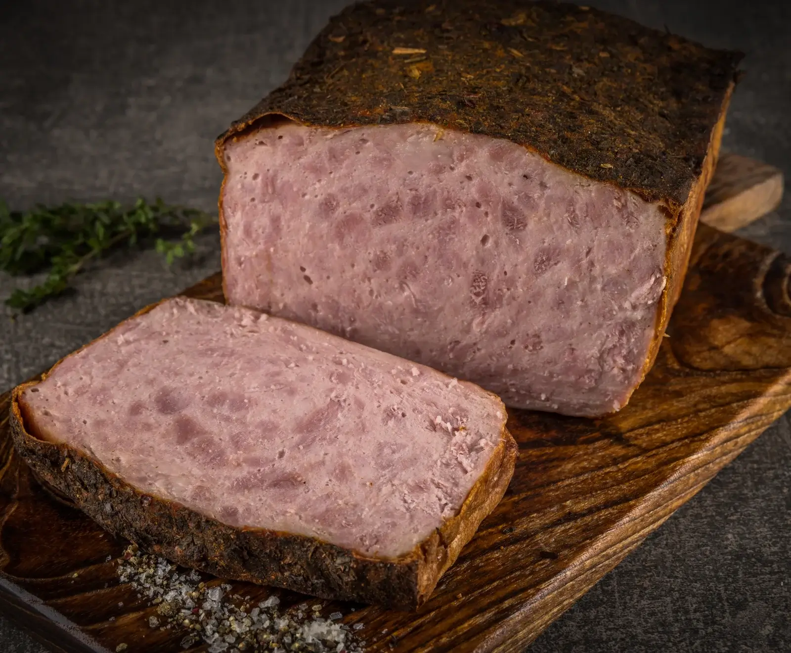 Angeschnittener Leberkäse mit grober, rosa Textur und dunkler Kräuterkruste auf einem Holzbrett, daneben grobes Salz und Pfeffer, mit Thymian im Hintergrund.