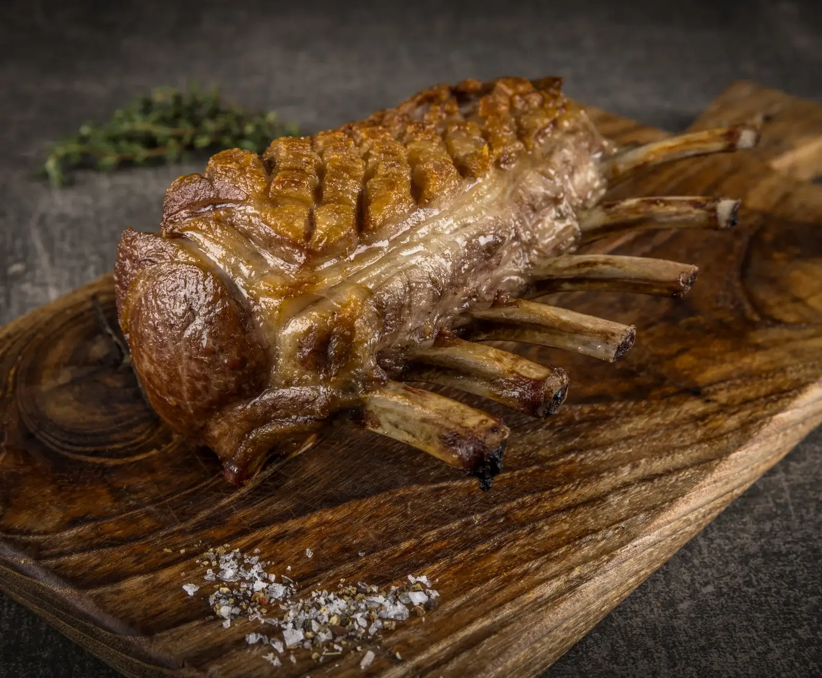 Ein perfekt gebratenes Lammkarree mit goldbrauner Kruste und sichtbaren Knochen liegt auf einem rustikalen Holzbrett. Grobes Meersalz und Pfeffer sind daneben gestreut, im Hintergrund ist Thymian zu sehen.