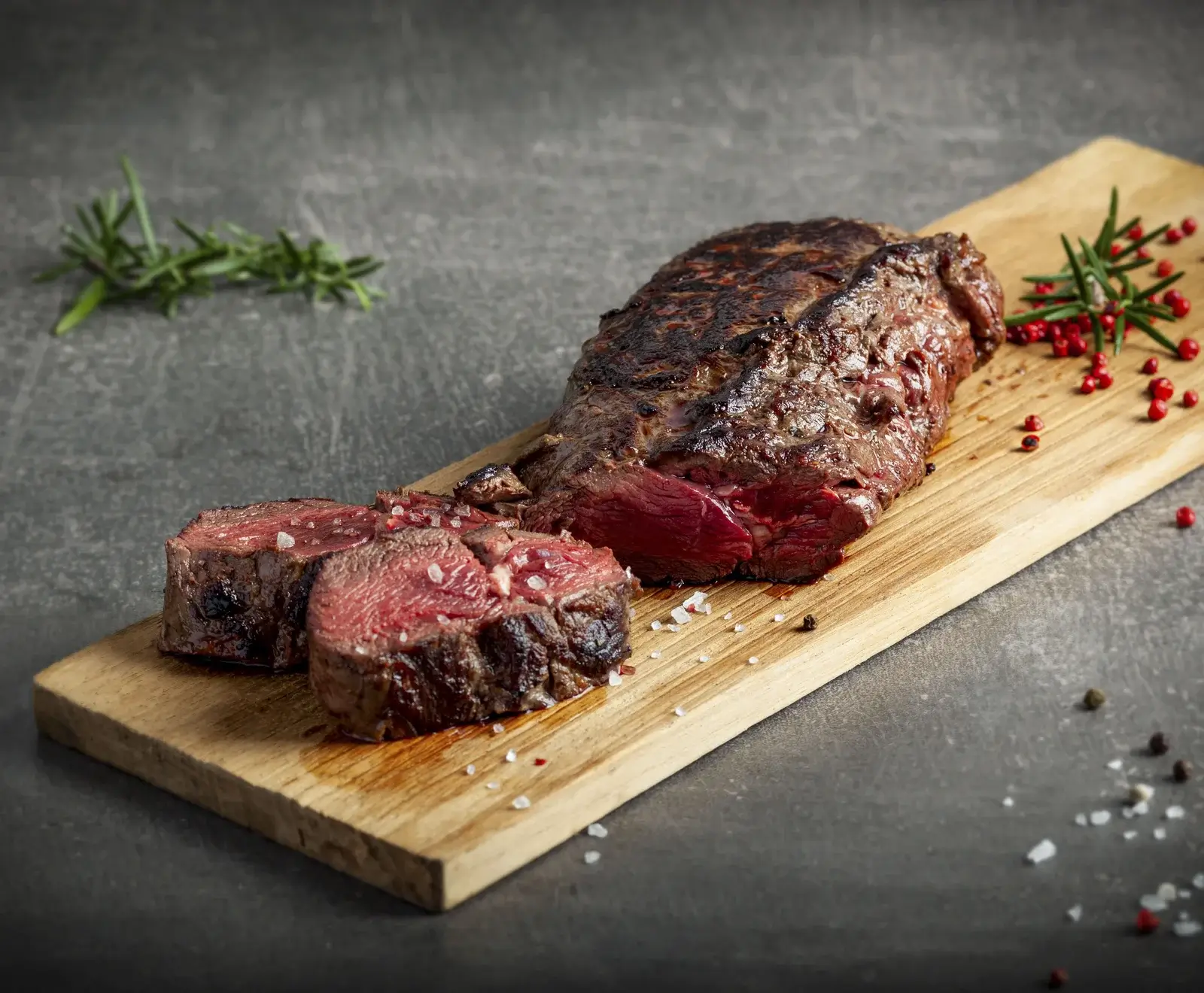 Ein perfekt gebratenes, teilweise aufgeschnittenes Steak auf einem Holzbrett, medium-rare mit grobem Salz, Rosmarin und roten Pfefferkörnern auf dunklem Hintergrund.