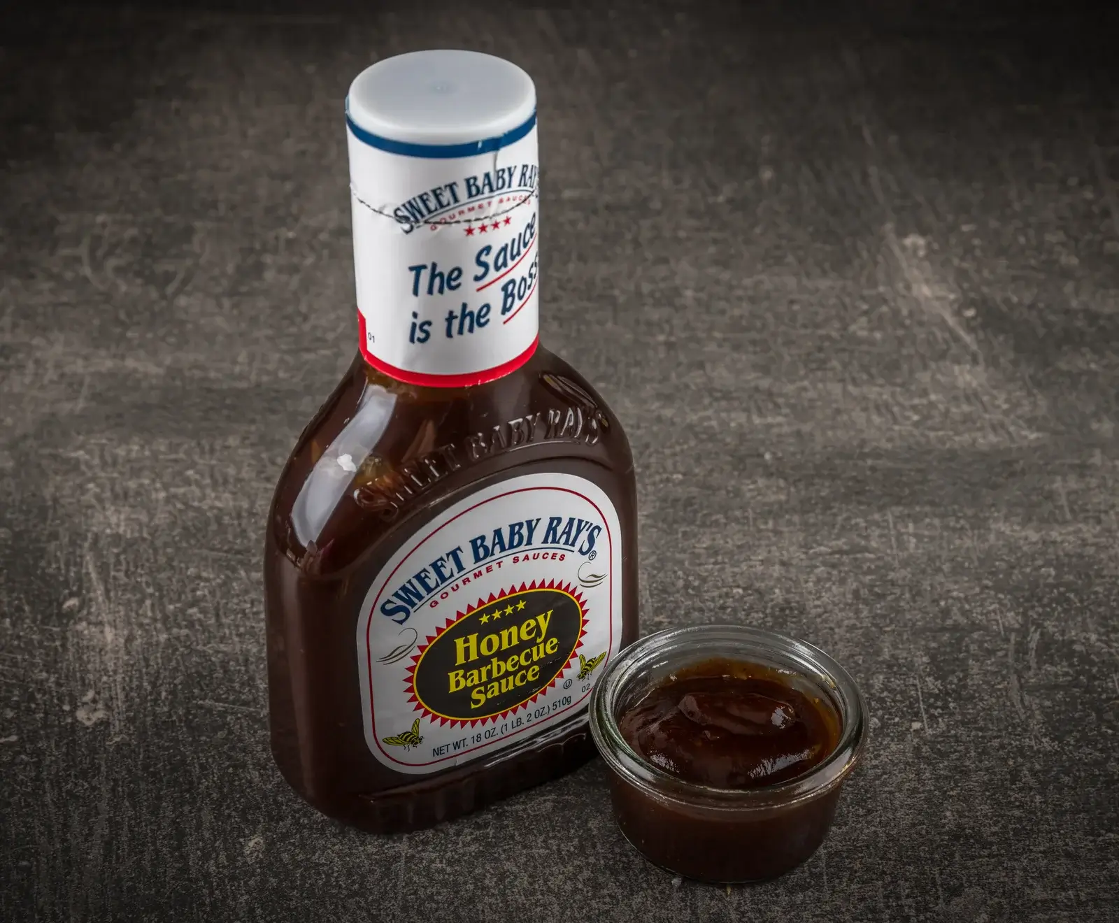 Eine Flasche Sweet Baby Ray's Honey Barbecue Sauce steht neben einem kleinen Glas mit der dunklen Sauce auf einem dunklen, strukturierten Untergrund. Das Etikett ist deutlich sichtbar.