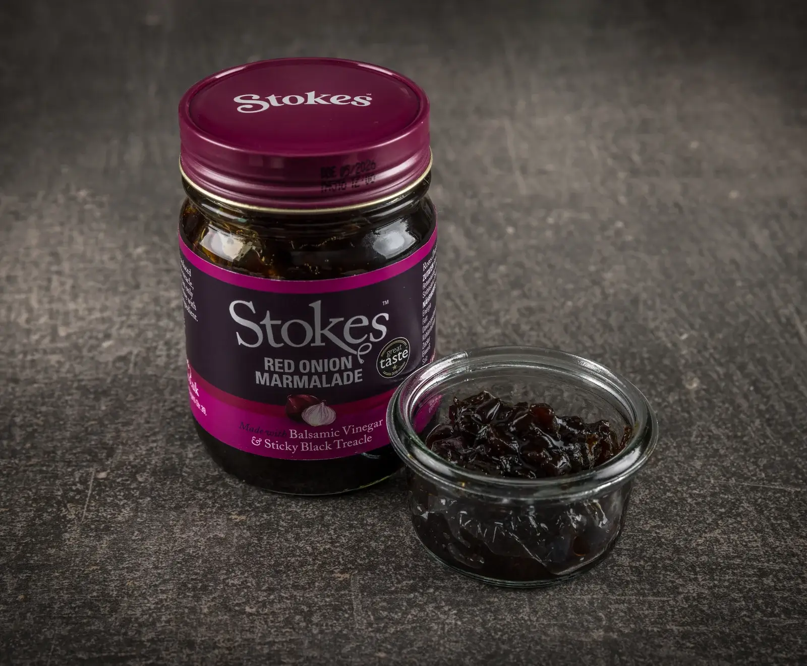 Ein Glas Stokes Rote Zwiebel Marmelade mit violetter Etikettierung und Deckel, daneben eine offene Glasschale mit der dunklen, stückigen Marmelade auf dunklem Untergrund.