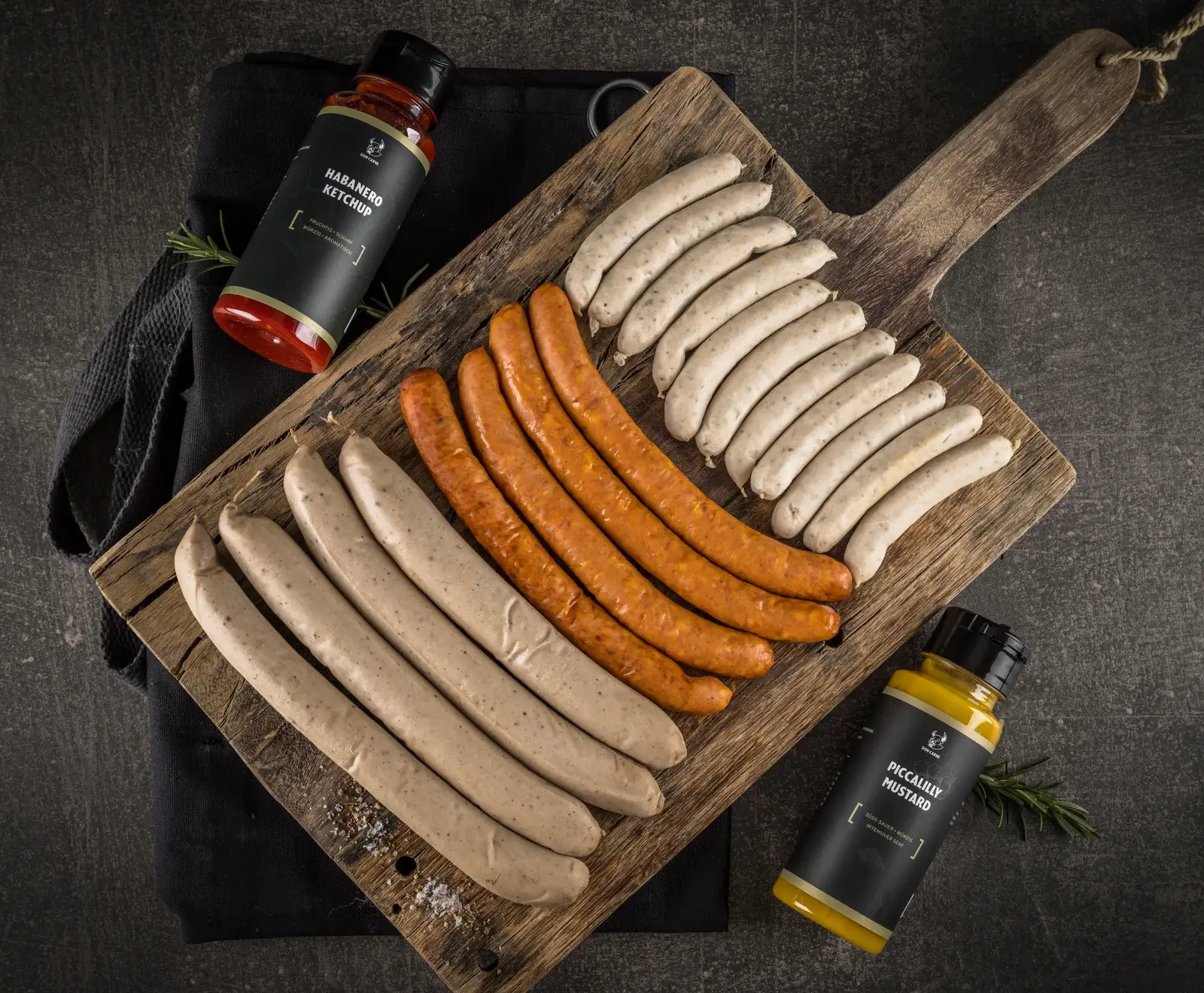 Holzbrett mit verschiedenen Würsten und zwei Flaschen Habanero-Ketchup auf dunklem Hintergrund.