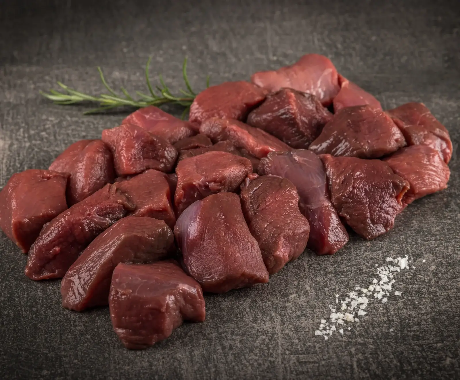 Ein Haufen roter, roher Wildfleischwürfel liegt auf einer dunklen, strukturierten Oberfläche. Im Hintergrund ist ein Rosmarinzweig zu sehen, im Vordergrund grobes Meersalz.