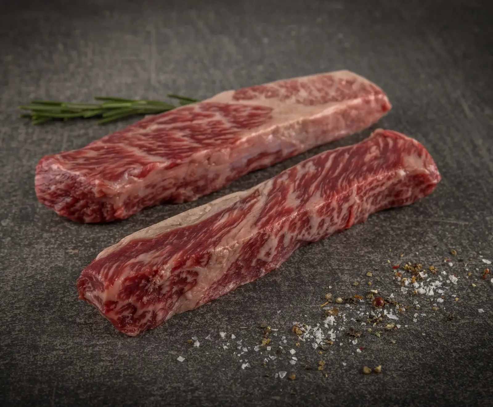 Zwei rohe, stark marmorierte Rindfleischstücke liegen auf einer dunklen Oberfläche. Im Hintergrund ist ein Rosmarinzweig, im Vordergrund grobes Salz und Pfeffer verstreut.