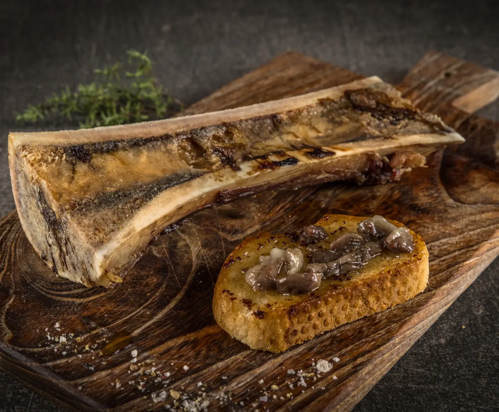 Gerösteter Markknochen und ein Toast mit Knochenmark auf einem Holzbrett, garniert mit Salz, Pfeffer und Thymian.