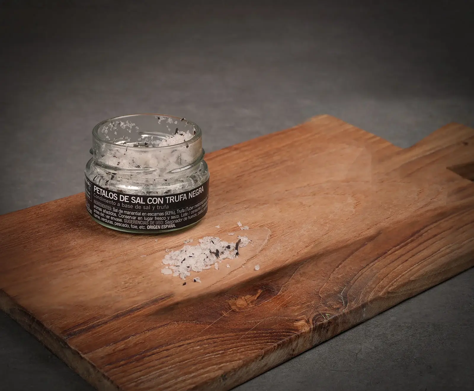 Ein offenes Glas "Petalos de Sal con Trufa Negra" (Salzflocken mit schwarzem Trüffel) steht auf einem Holzschneidebrett. Daneben liegt eine kleine Menge des Salzes verstreut.