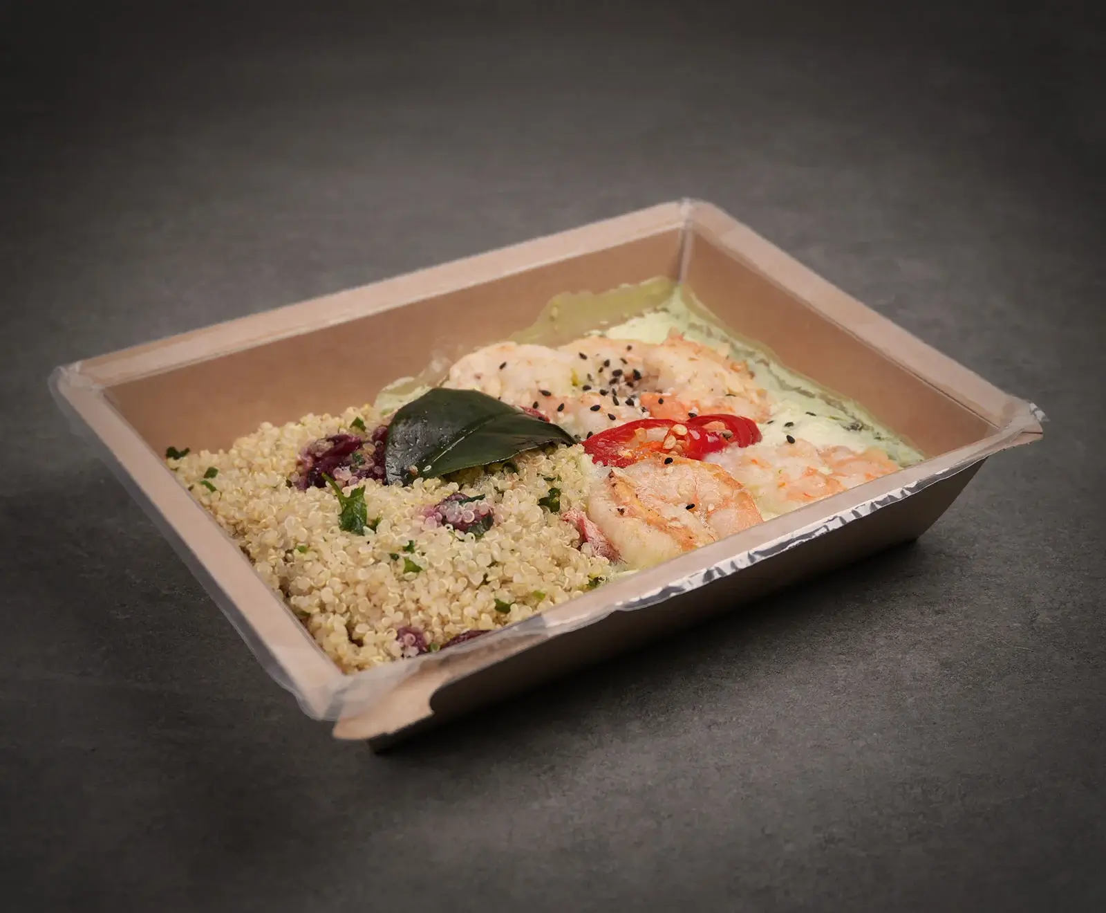 Eine Mahlzeit in einer braunen Pappschale mit Klarsichtfolie. Links Quinoa mit Kräutern und roten Körnern, rechts gekochte Garnelen in heller grüner Soße, garniert mit Chili, schwarzem Sesam und einem Blatt.