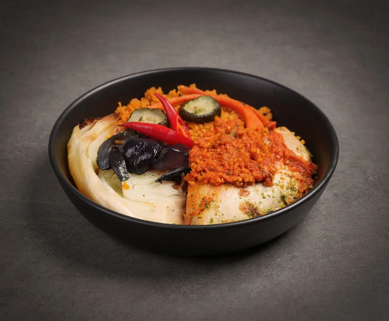 Eine schwarze Schale mit einem Gericht auf dunklem Hintergrund. Es enthält gekochte Weißkohlblätter, Auberginenstücke, orangefarbenen Bulgur, rote Chilischoten, eingelegte Gurken und ein Fischfilet mit roter Sauce.