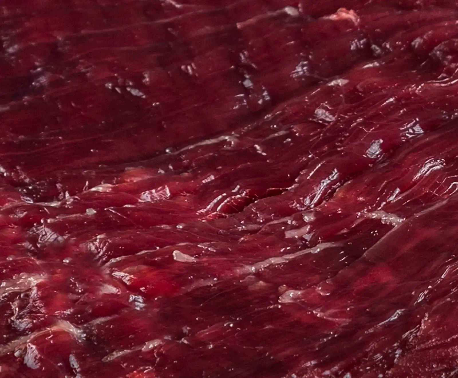 Nahaufnahme von dunkelrotem, rohem Fleisch mit sichtbaren Muskelfasern und feinen weißen Fett- oder Bindegewebsstreifen. Die Oberfläche glänzt leicht.
