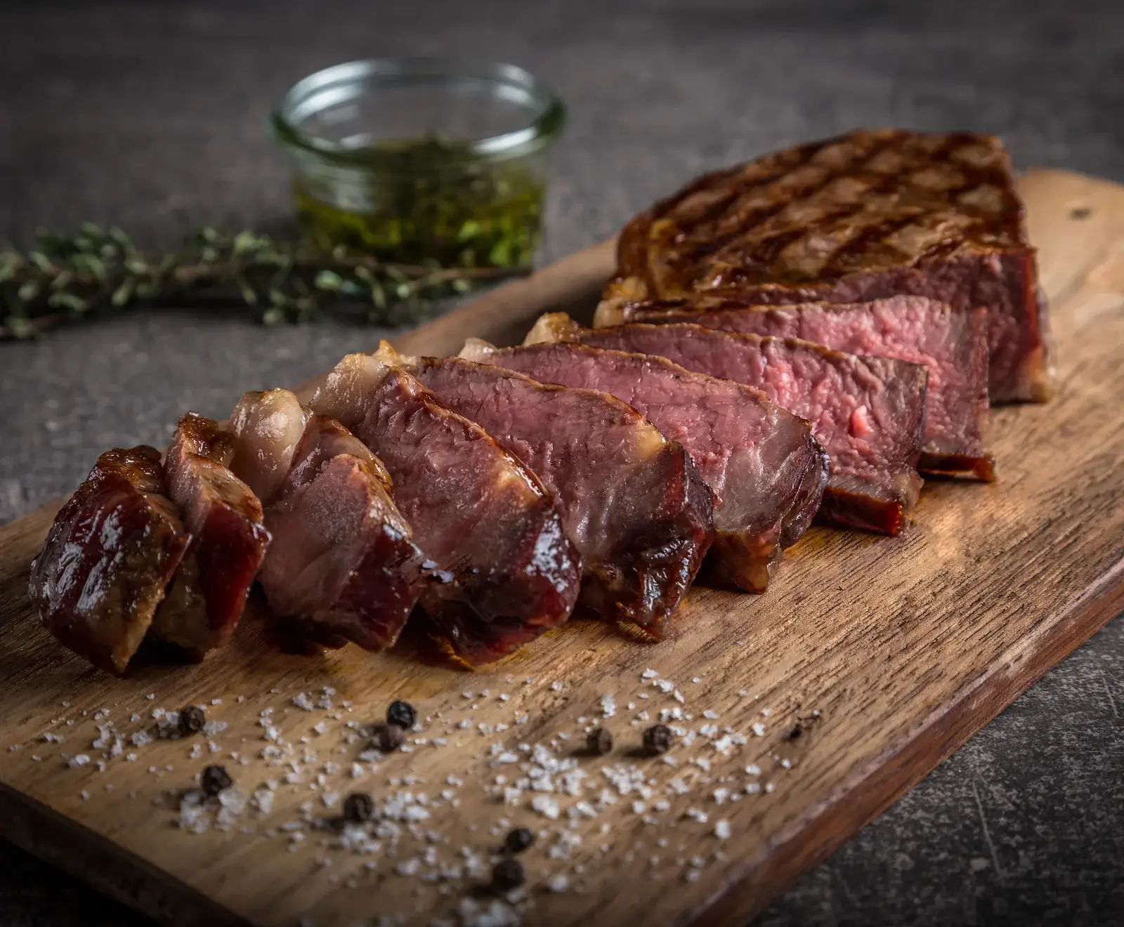 Ein aufgeschnittenes, medium-rare gegrilltes Steak auf einem Holzbrett, bestreut mit grobem Salz und Pfeffer. Im Hintergrund ein Glas mit Kräutersauce und Thymianzweigen.