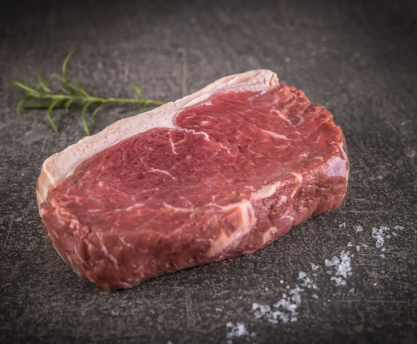 Ein rohes Rindersteak mit Fettrand liegt auf einer dunklen Steinplatte, begleitet von einem Rosmarinzweig und grobem Meersalz.