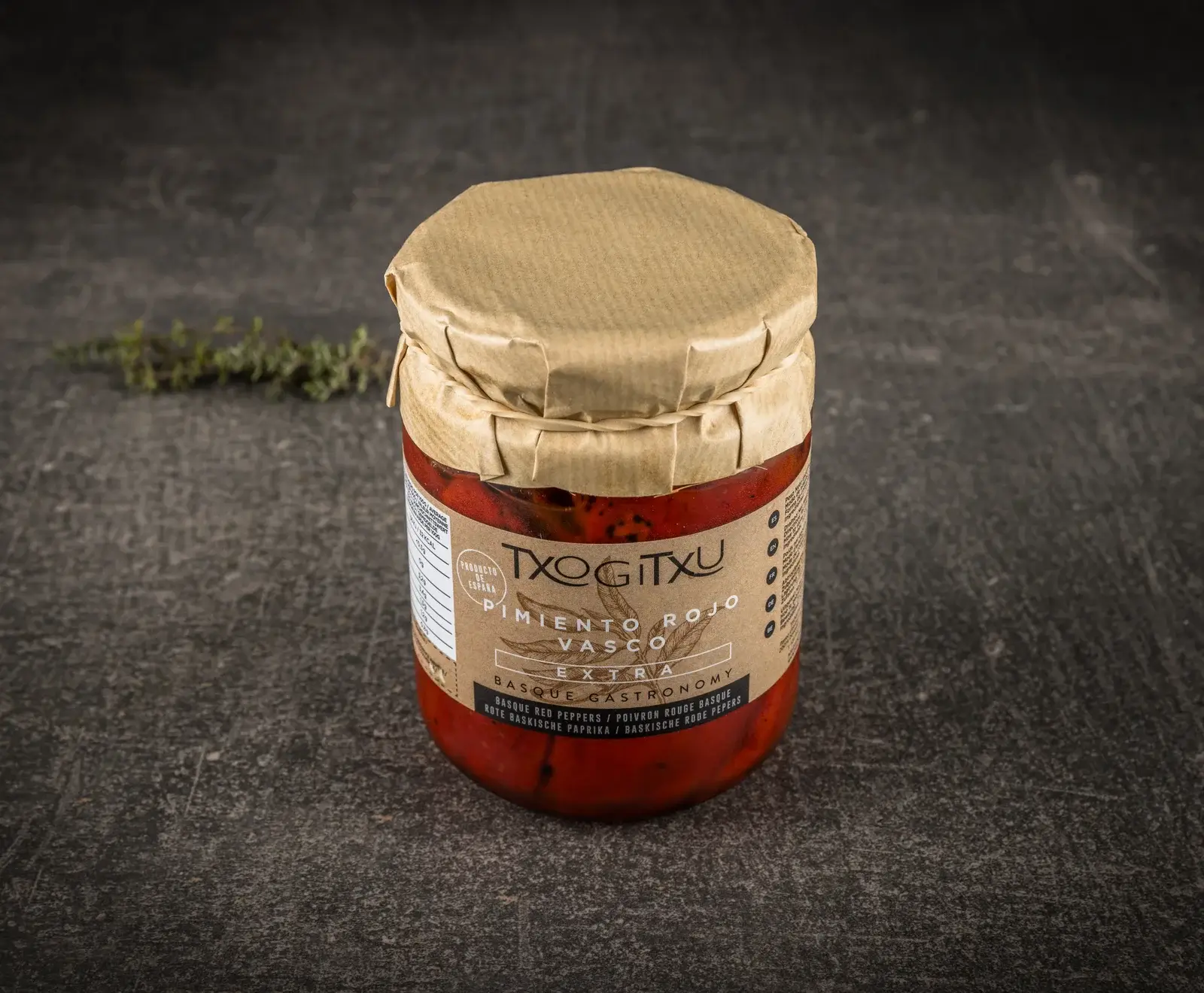 Ein Glas TXOGITXU Pimiento Rojo Vasco Extra (Baskische Rote Paprika) mit braunem Papierdeckel, gefüllt mit roter Paprikapaste, auf dunklem Untergrund. Ein Thymianzweig liegt im Hintergrund.
