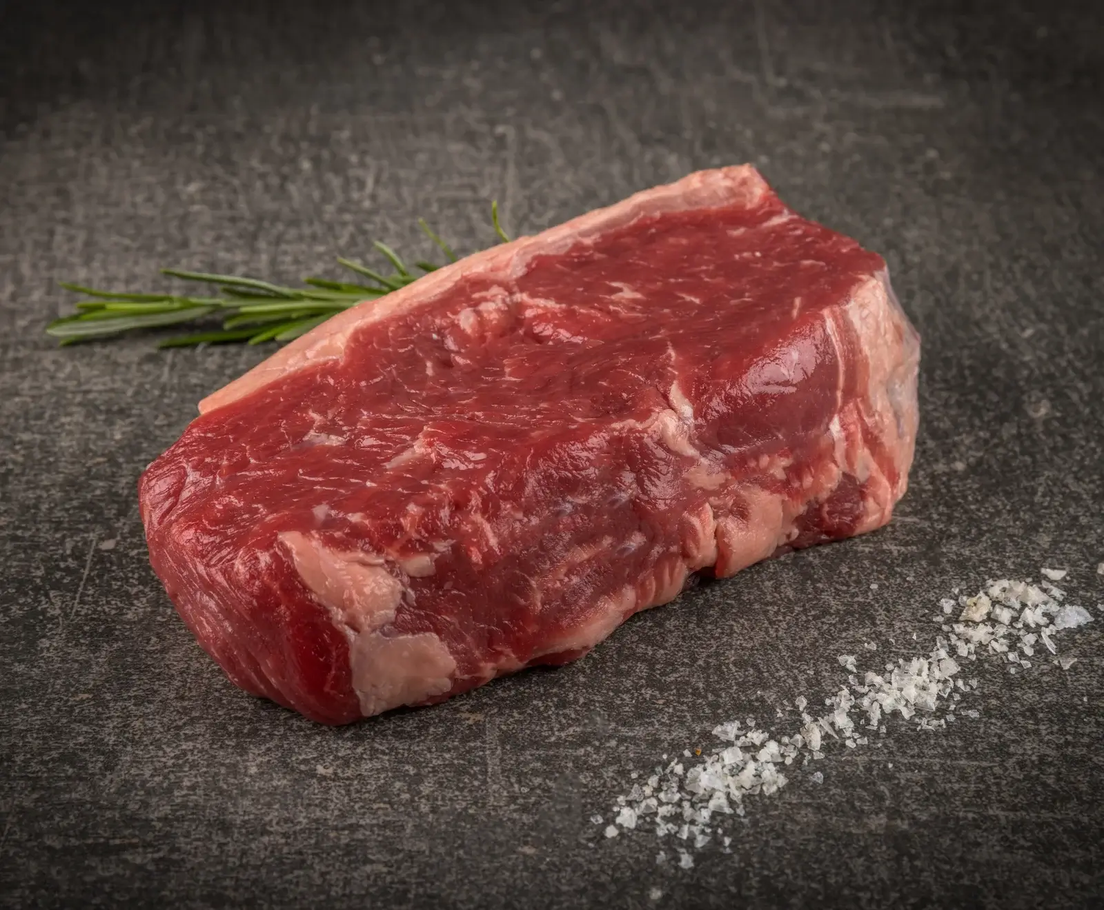 Ein rohes, rotes Rindersteak mit einem weißen Fettdeckel liegt auf einer dunklen, strukturierten Oberfläche. Im Hintergrund ist ein Rosmarinzweig, im Vordergrund eine Spur grobes Meersalz.