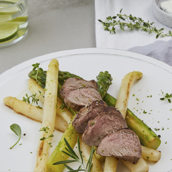 Nahaufnahme eines Tellers mit rosa gebratenem Fleisch, in Scheiben geschnitten, auf weißem und grünem Spargel angerichtet, garniert mit frischen Kräutern wie Thymian und Rosmarin.