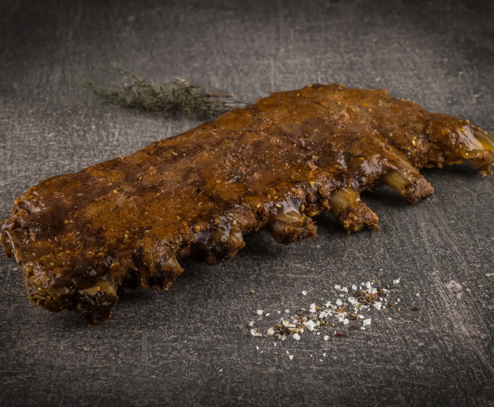 Gebratene Rippchen vom Rind auf grauem Hintergrund, daneben verstreut grobes Salz und ein Zweig Rosmarin