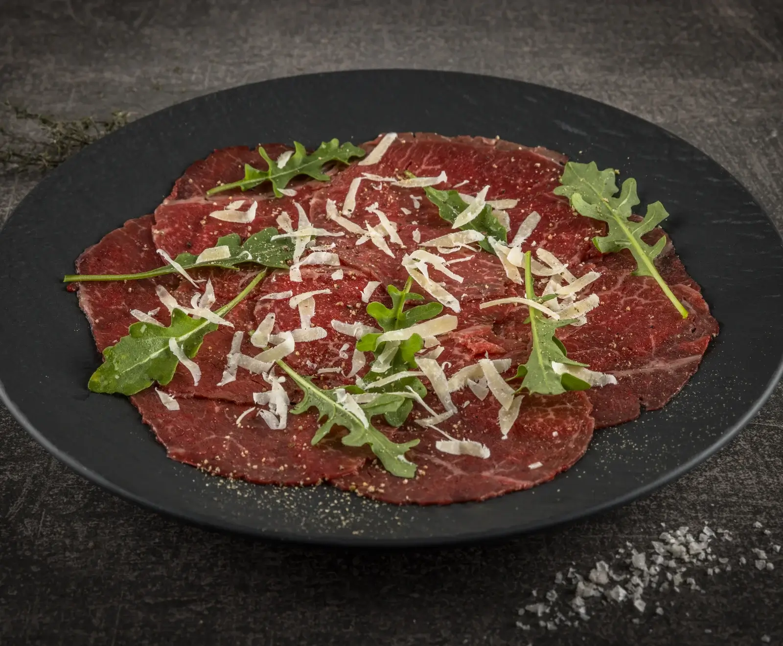 Nahaufnahme von Rindercarpaccio auf einem schwarzen Teller, garniert mit geriebenem Parmesan, frischem Rucola und Pfeffer. Der dunkle Hintergrund zeigt grobes Salz.