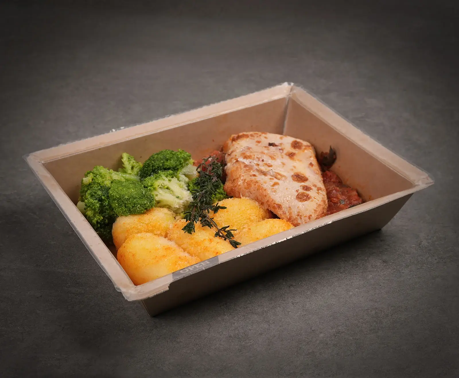 Ein Fertiggericht in einer braunen Pappschale mit transparentem Deckel. Es enthält ein überbackenes Hähnchenfilet, goldgelbe Kartoffeln, frischen Brokkoli und Tomatensauce, garniert mit Thymian, auf dunklem Untergrund.