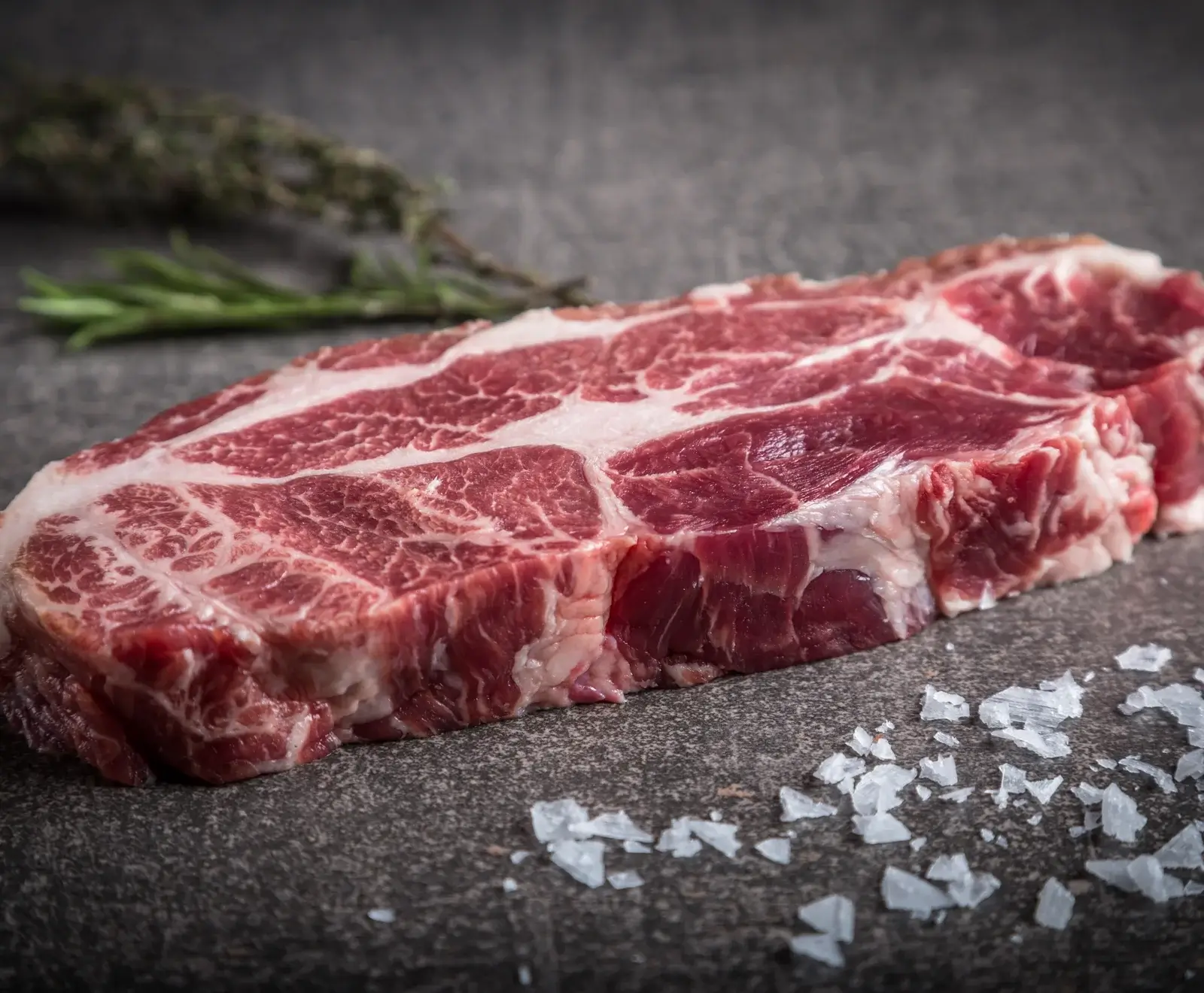 Rohes Steak mit Meersalz auf dunklem Hintergrund, daneben ein Zweig Rosmarin