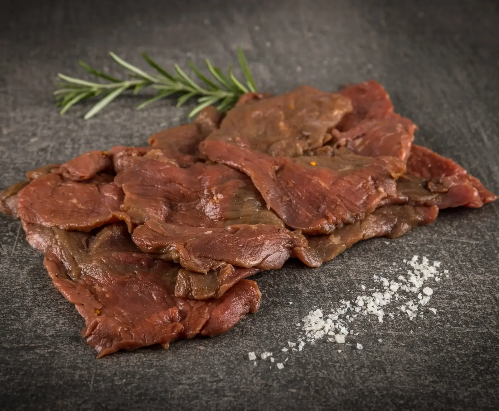 Dünn geschnittenes, rötlich-braunes Fleisch auf einer dunklen Steinplatte, garniert mit einem Rosmarinzweig und grobem Meersalz.
