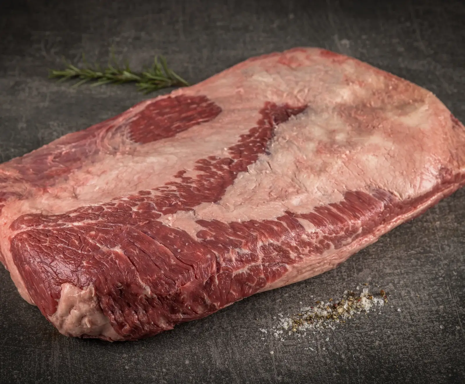 Ein großes, rohes Rinderbruststück (Brisket) mit deutlicher Marmorierung und Fettschicht liegt auf dunklem Untergrund. Im Hintergrund ein Rosmarinzweig, im Vordergrund Salz und Pfeffer.