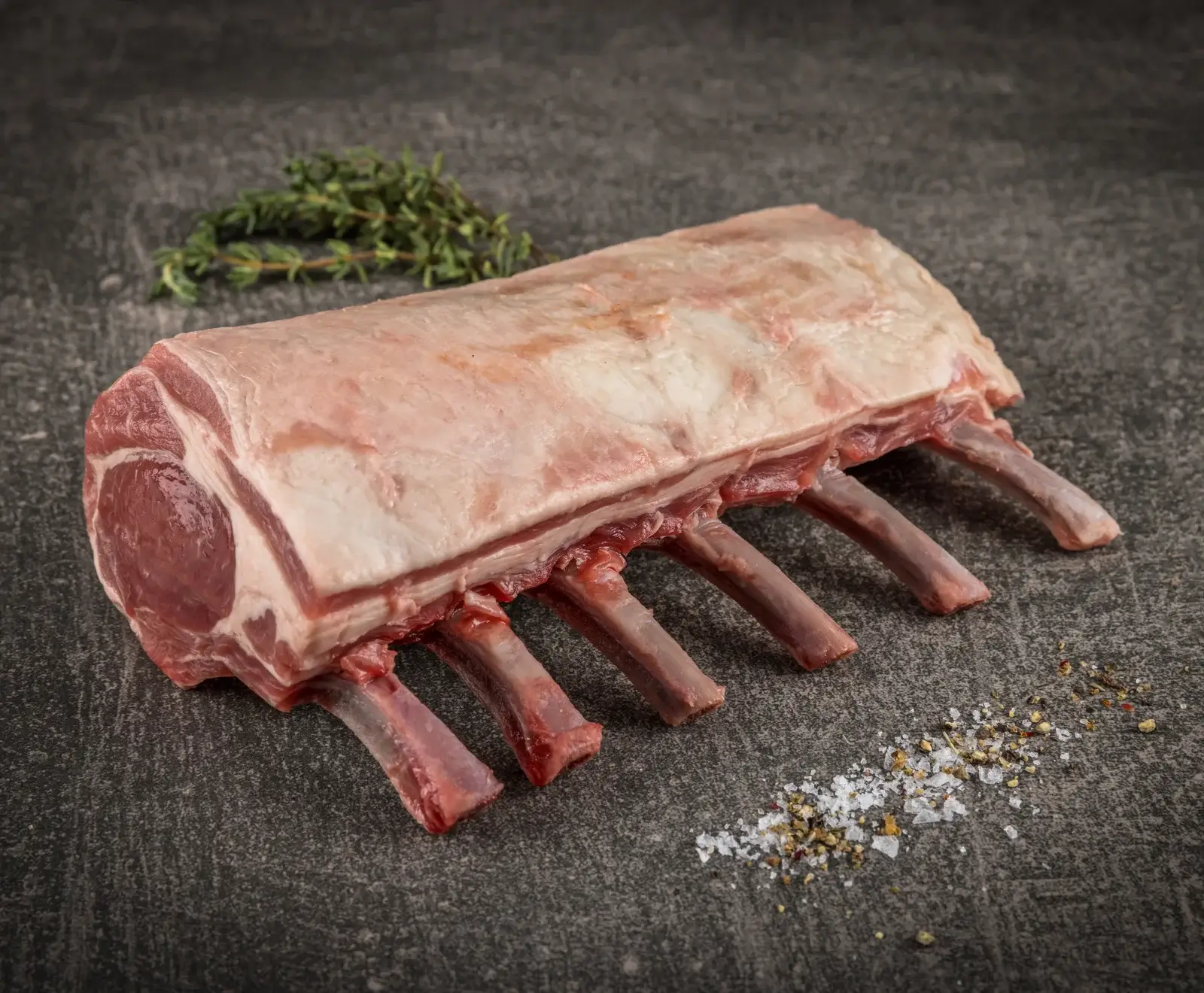 Rohes Lammkarree mit sichtbaren Rippen und Fettschicht, auf dunklem Untergrund. Daneben liegen frischer Thymian, grobes Salz und Pfeffer.