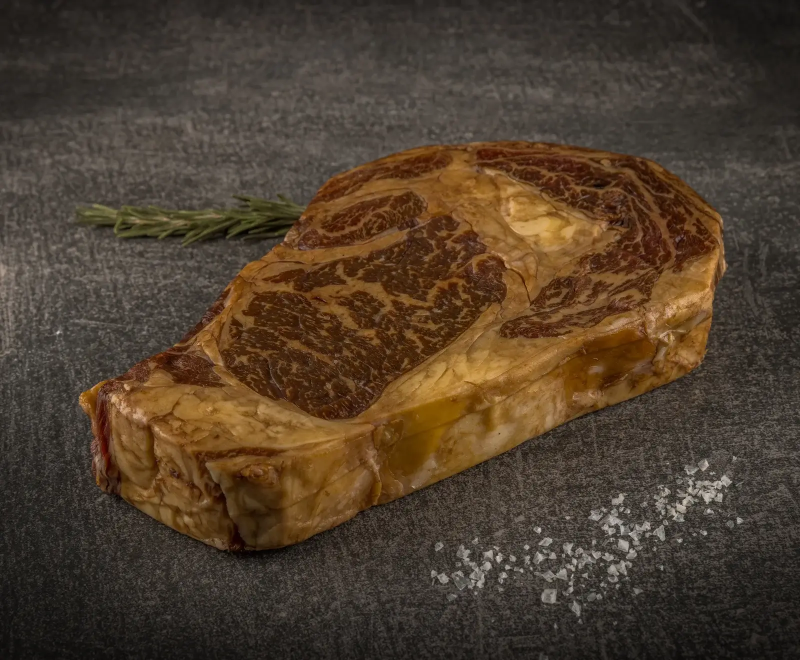Gegrilltes Ribeye Steak mit Rosmarin auf einer dunklen Steinplatte mit Salz daneben.