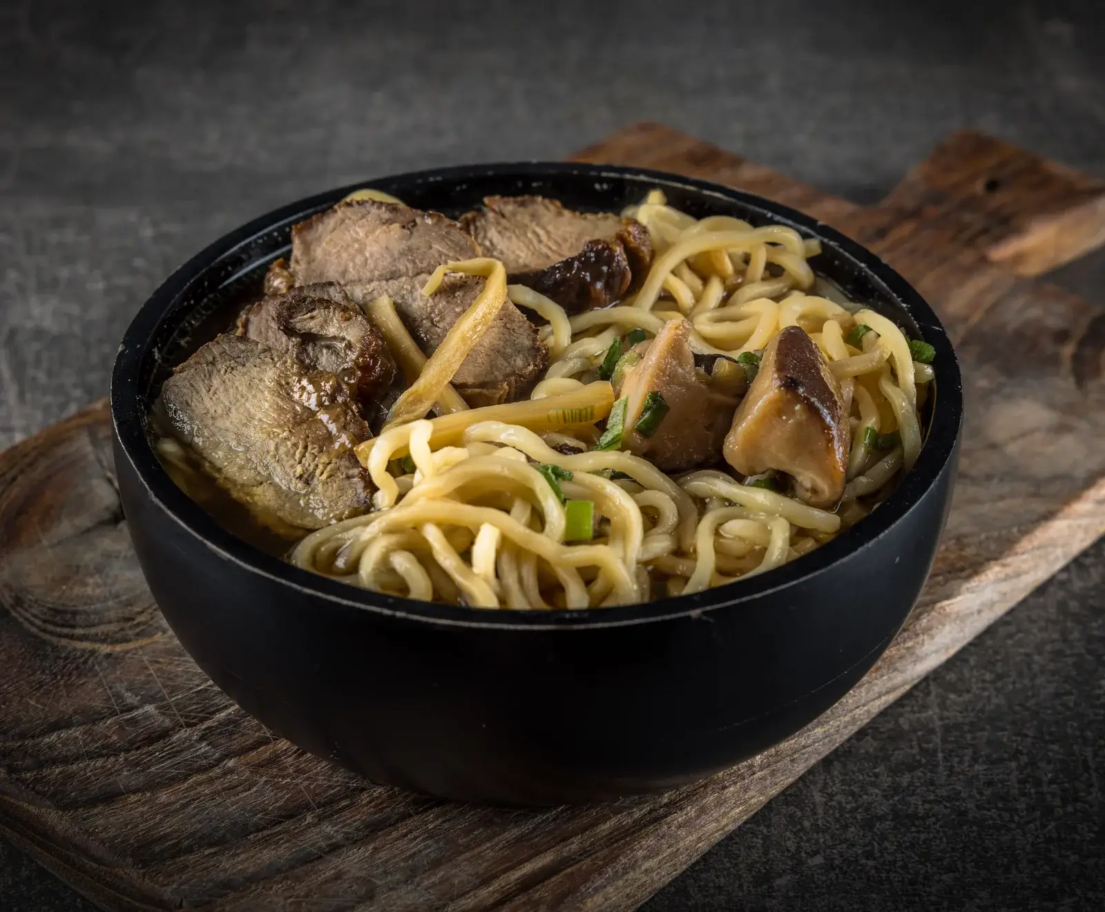 Eine schwarze Schale mit Ramen-Nudeln, Rindfleischscheiben, Pilzen, Bambussprossen und Frühlingszwiebeln in Brühe, auf einem Holzbrett vor dunklem Hintergrund.