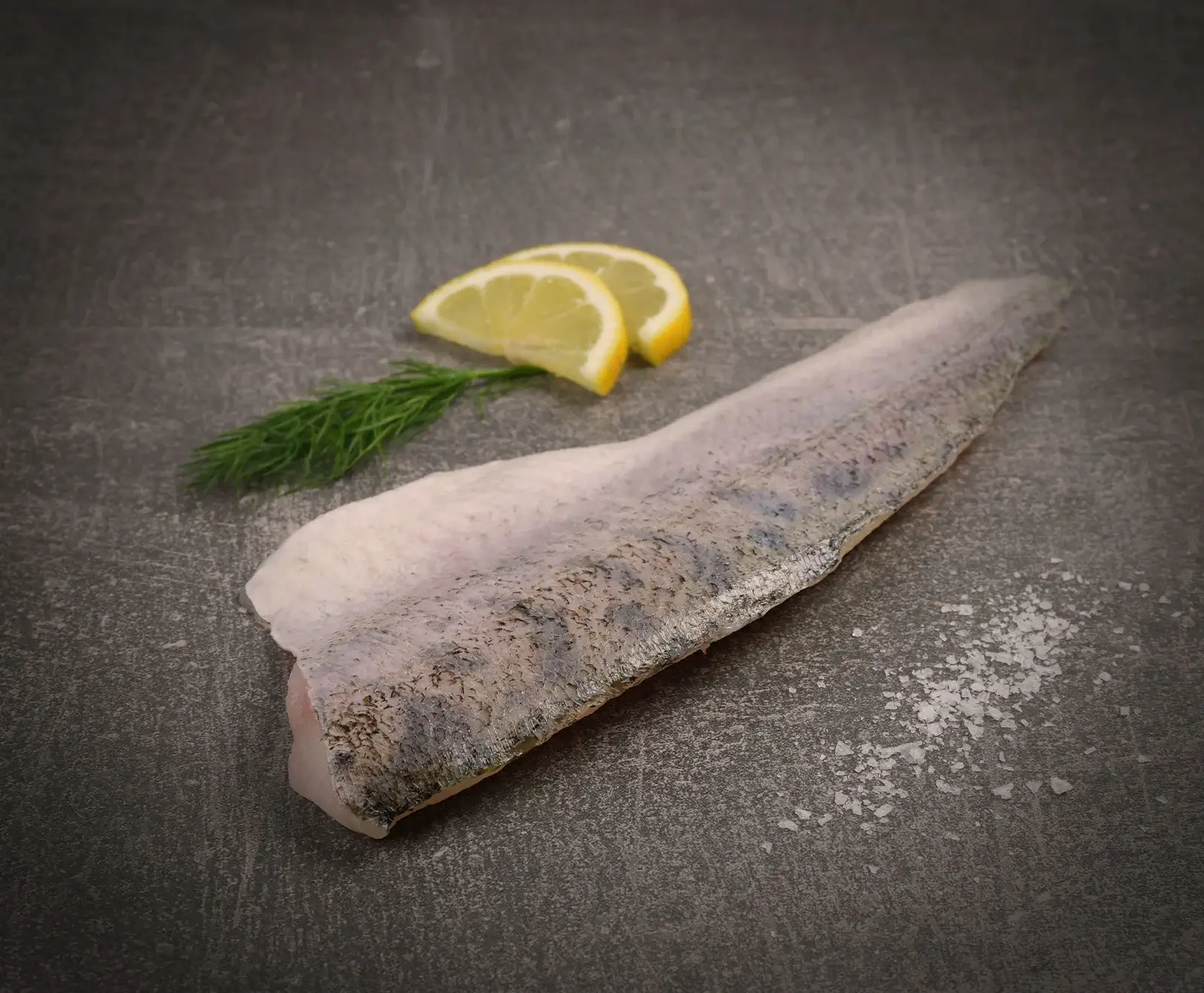 Ein rohes Fischfilet mit Haut liegt auf einer dunklen, strukturierten Oberfläche. Daneben sind zwei Zitronenspalten, ein Dillzweig und grobes Meersalz verstreut.