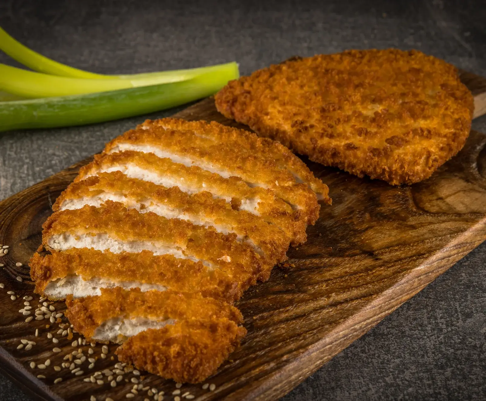 Zwei goldbraun panierte Hähnchenschnitzel auf einem Holzbrett. Eines ist ganz, das andere in Streifen geschnitten. Grüne Lauchzwiebeln und Sesamsamen sind im Hintergrund verstreut.