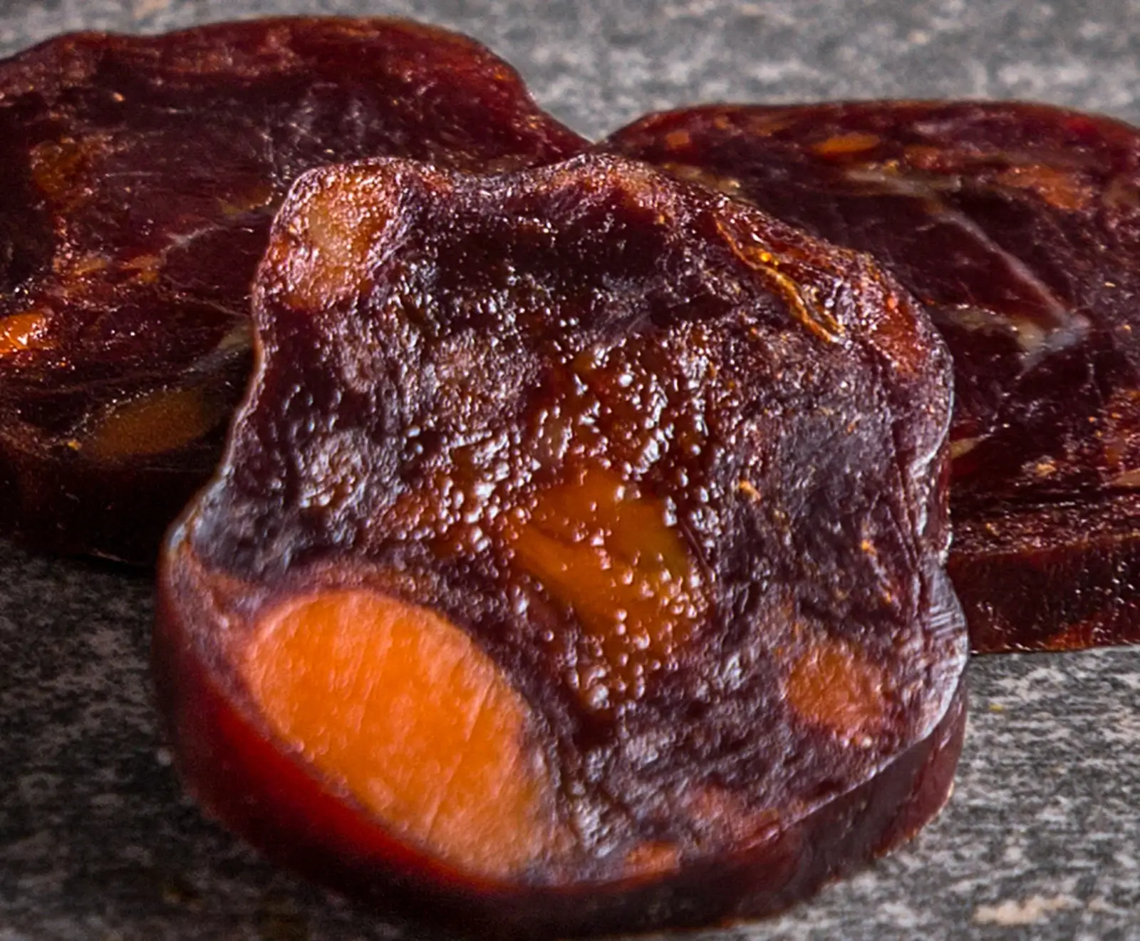 Nahaufnahme von drei glänzenden, dunkelbraunen Trockenfrüchten mit orangefarbenen Einschlüssen, die auf einer grauen, gesprenkelten Oberfläche liegen.