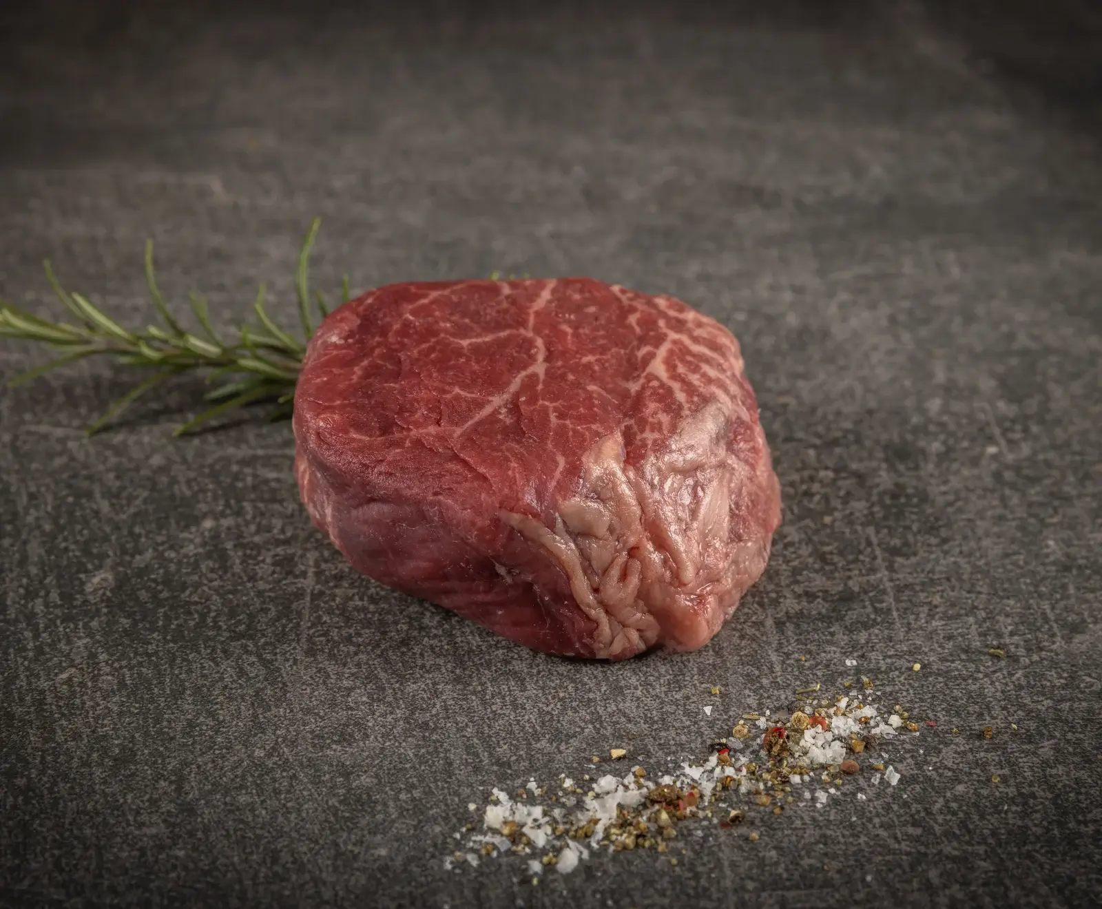 Ein rohes, marmoriertes Rinderfilet liegt auf einer dunklen Steinplatte. Daneben ein Rosmarinzweig und eine Prise grobes Salz mit Pfefferkörnern.