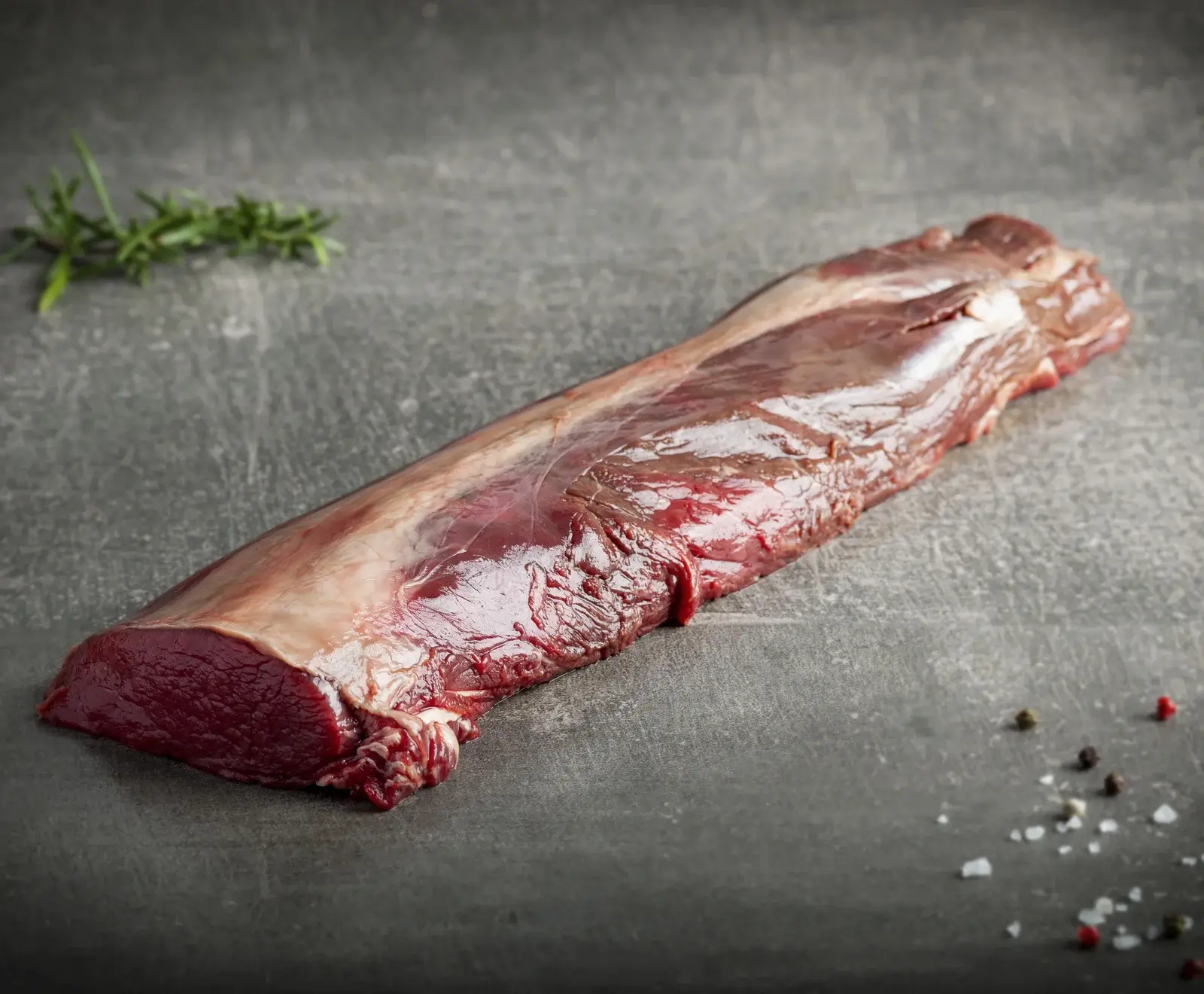 Ein rohes, dunkelrotes Wildfilet liegt auf einer dunklen Steinplatte, umgeben von Rosmarinzweigen, bunten Pfefferkörnern und grobem Salz. Die Oberfläche des Fleisches glänzt leicht.
