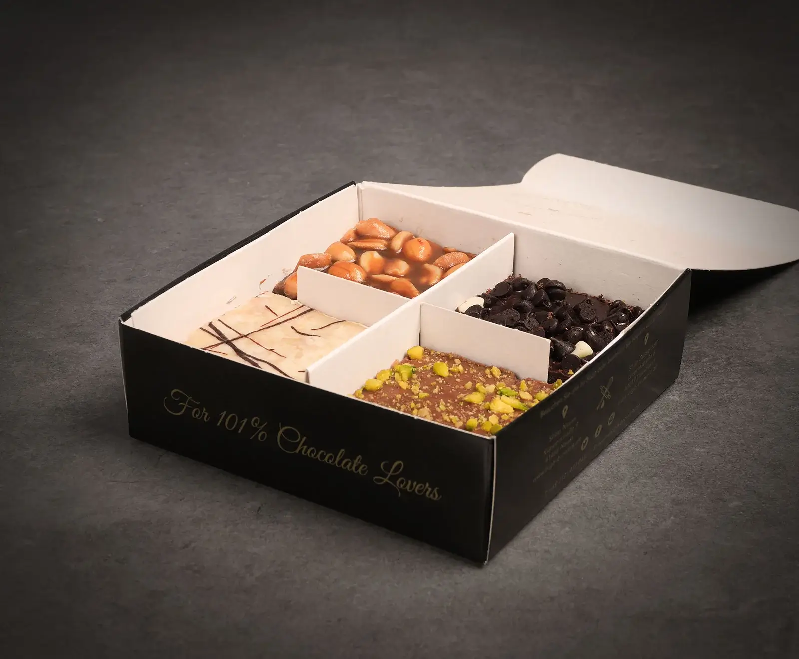 Eine offene schwarze Schachtel mit vier Fächern, gefüllt mit verschiedenen Schokoladen- und Nuss-Spezialitäten. Zu sehen sind Karamellnüsse, Schokochips, weiße Schokolade und Pistazien-Schokolade. Die Box trägt die Aufschrift "For 101% Chocolate Lov